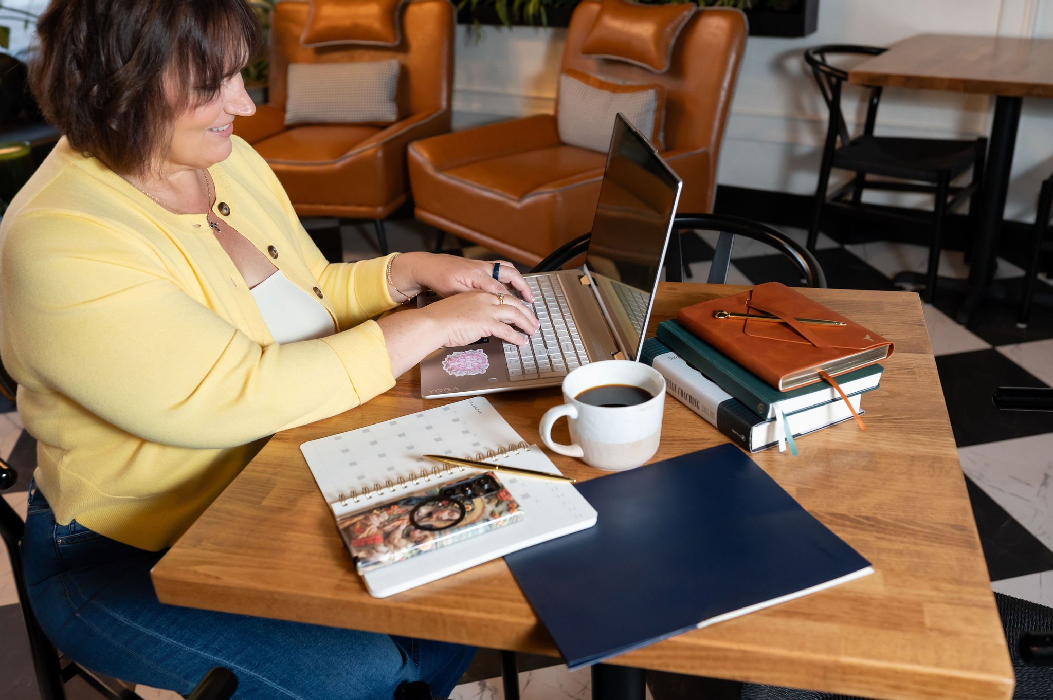 Woman in yellow sweater sitting at a wooden table, typing on a laptop, with notebooks, a cup of coffee, and a planner in front of her, in a cozy cafe setting.