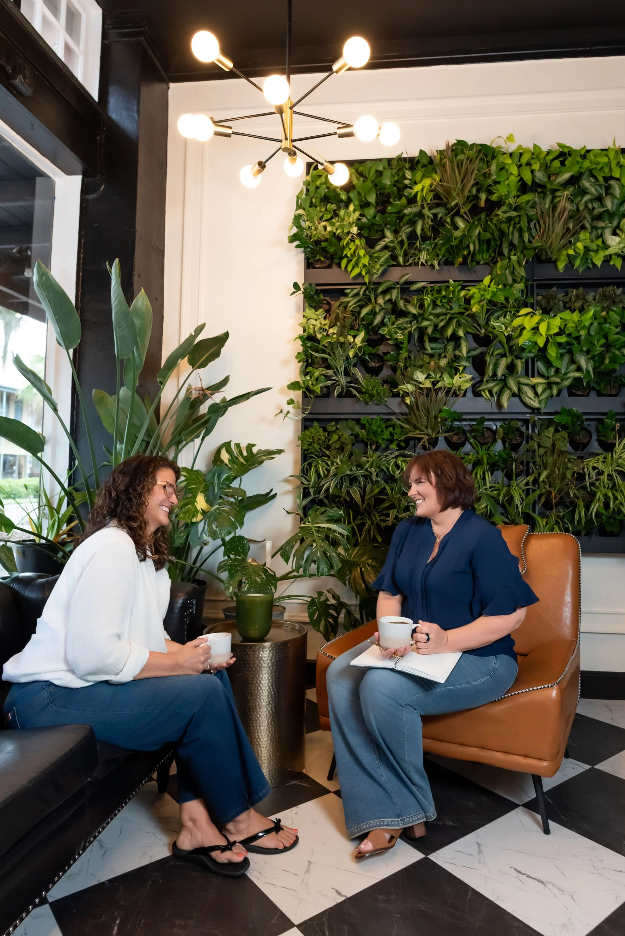 Two women sitting in a cafe, smiling and talking. One woman has curly hair and glasses, holding a mug; the other has short hair, wearing a blue shirt, holding a mug, with a notebook on her lap. The cafe has a green plant wall, black and white checkered floor, and a modern chandelier.