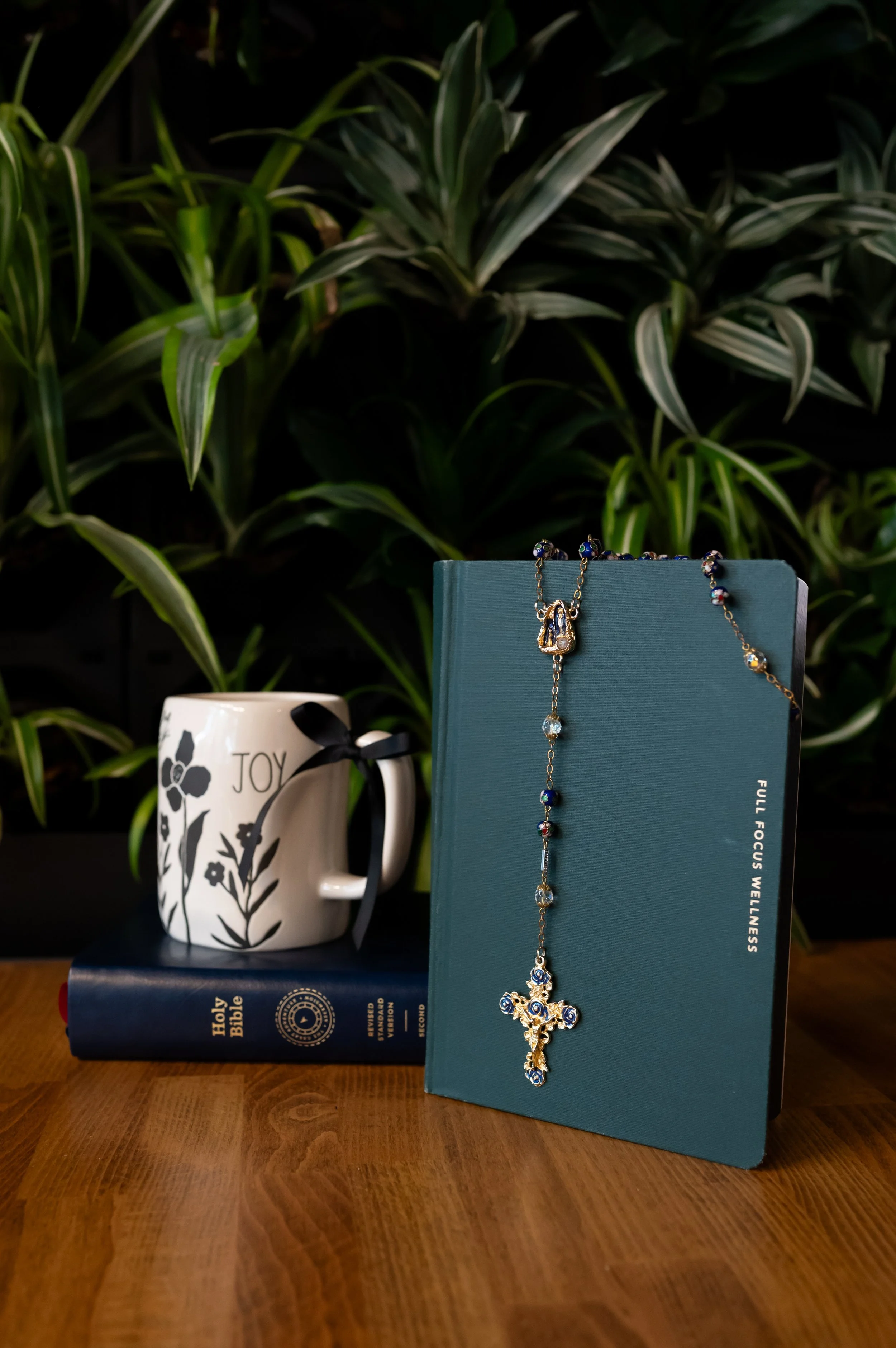 A white mug with floral design and the word 'Joy' printed on it, a blue book with religious text on the cover, a jewelry necklace with a floral cross pendant draped over the book, a green plant in the background, and a wooden table surface.