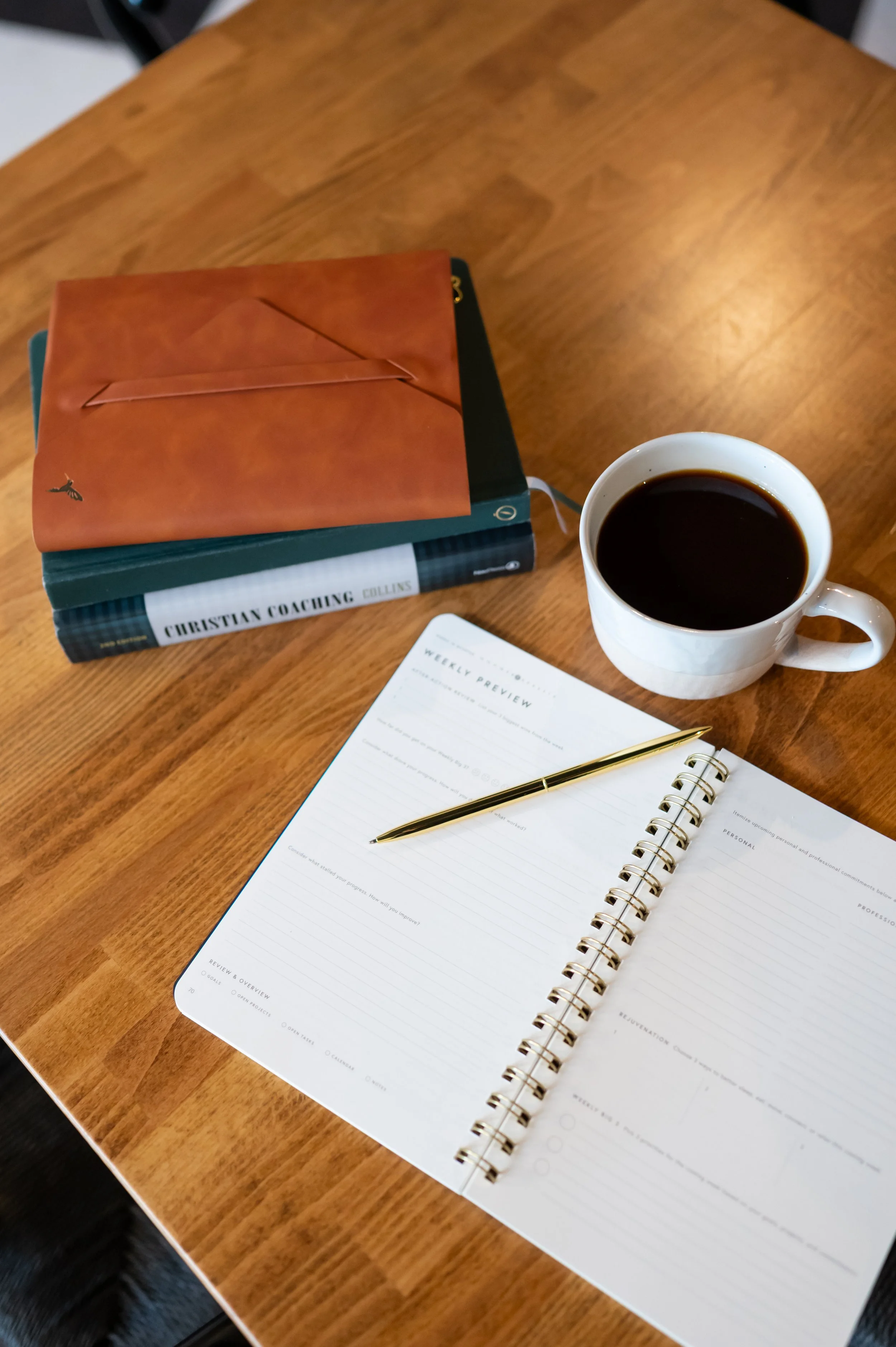 Open planner with weekly preview pages, a gold pen, a cup of black coffee, and books on a wooden table.