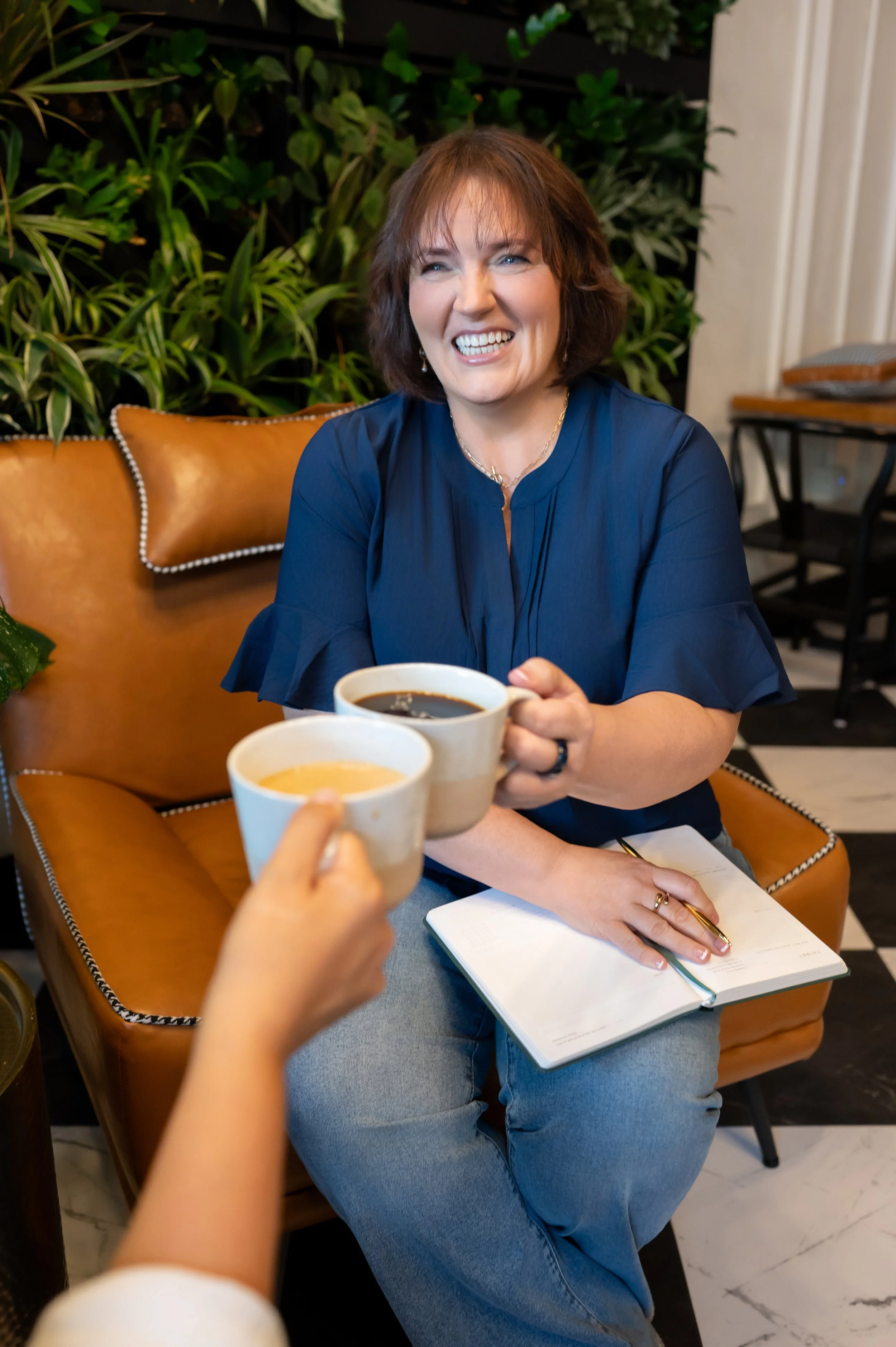A woman smiling and holding a cup of coffee, sitting at a table in a cozy cafe. Two more cups of coffee are being offered to her, and she has a notebook and pen in her lap. There are green plants and comfortable seating in the background.