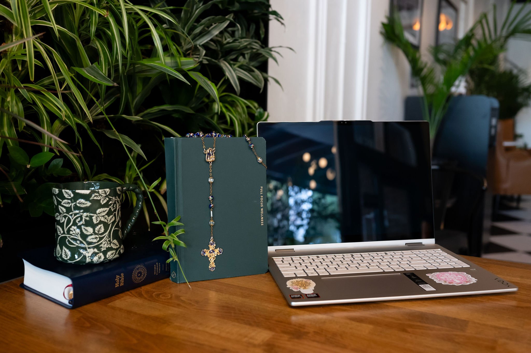 A desk with a laptop, a green book with religious text, a decorative mug, hanging jewelry, a smartphone, and several green houseplants.