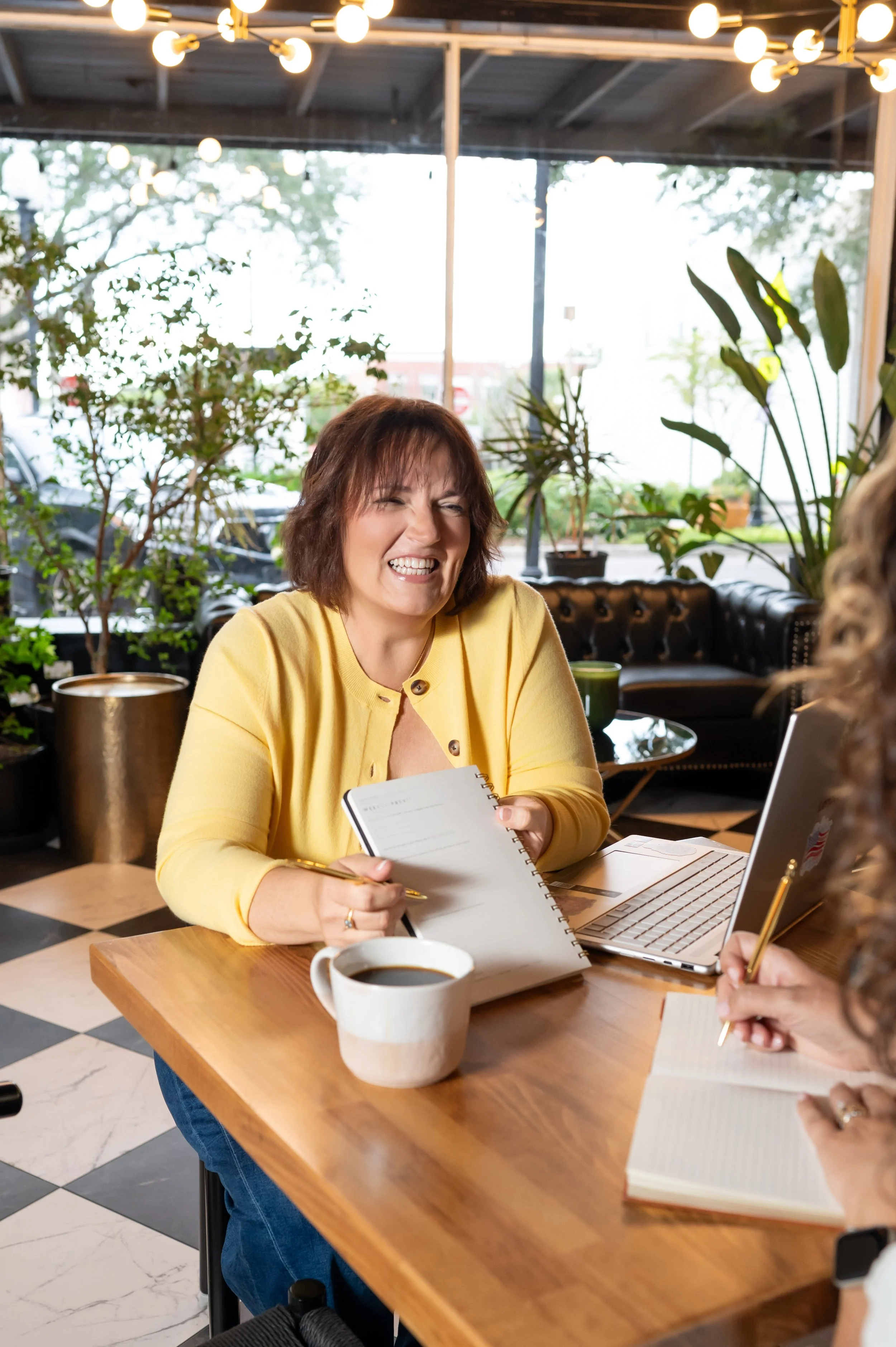 A woman in a yellow top sitting at a wooden table in a cafe, smiling and holding a notebook, with a cup of coffee, a laptop, and people taking notes nearby, surrounded by plants and string lights.