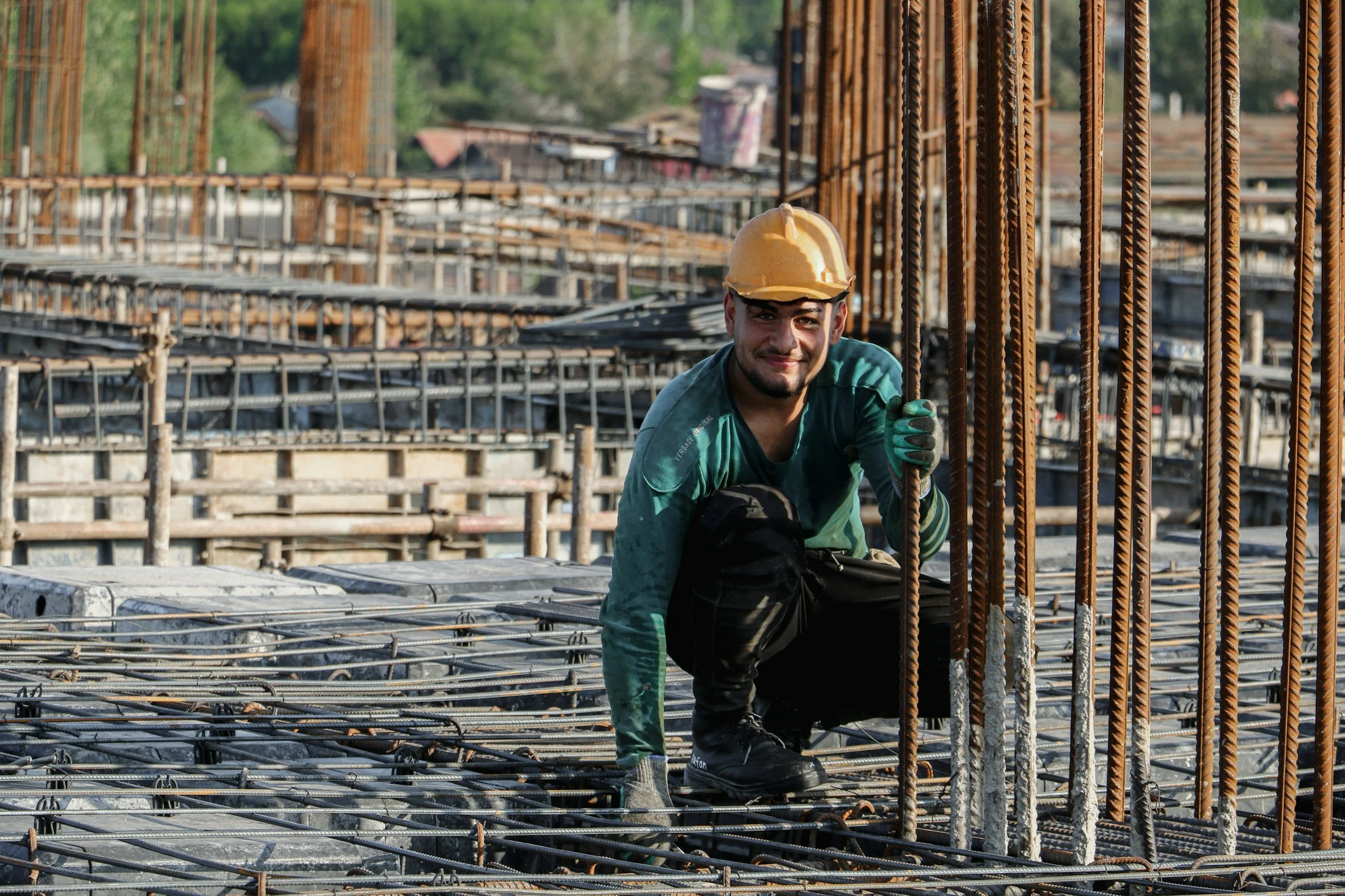 Construction worker wearing a yellow safety helmet and green gloves, smiling, among steel rebar on a building site with partially constructed framework in the background.