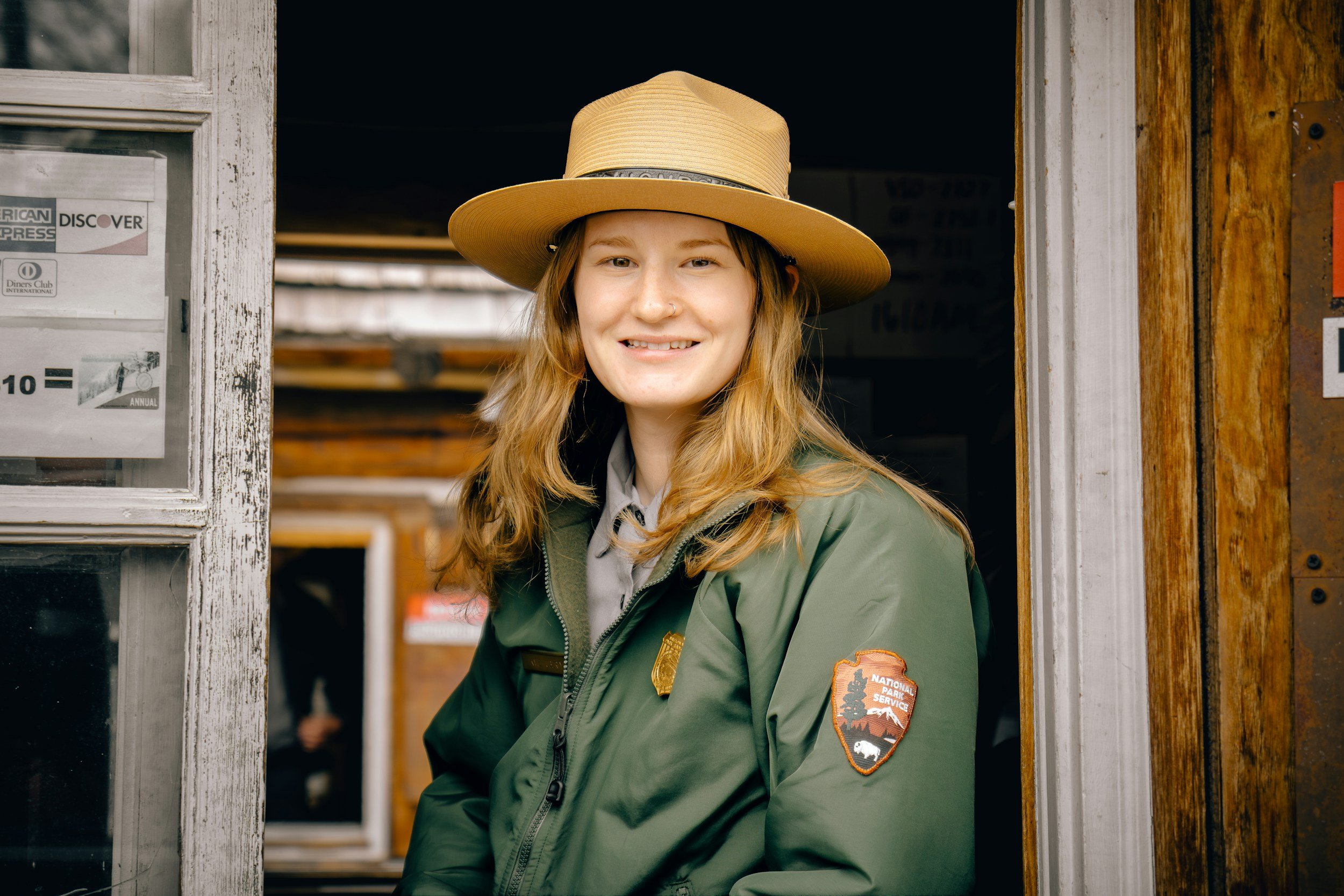 A young woman with long, wavy red hair, smiling, standing at a rustic doorway wearing a tan wide-brimmed hat and a green jacket with patches, including one for National Park Service.