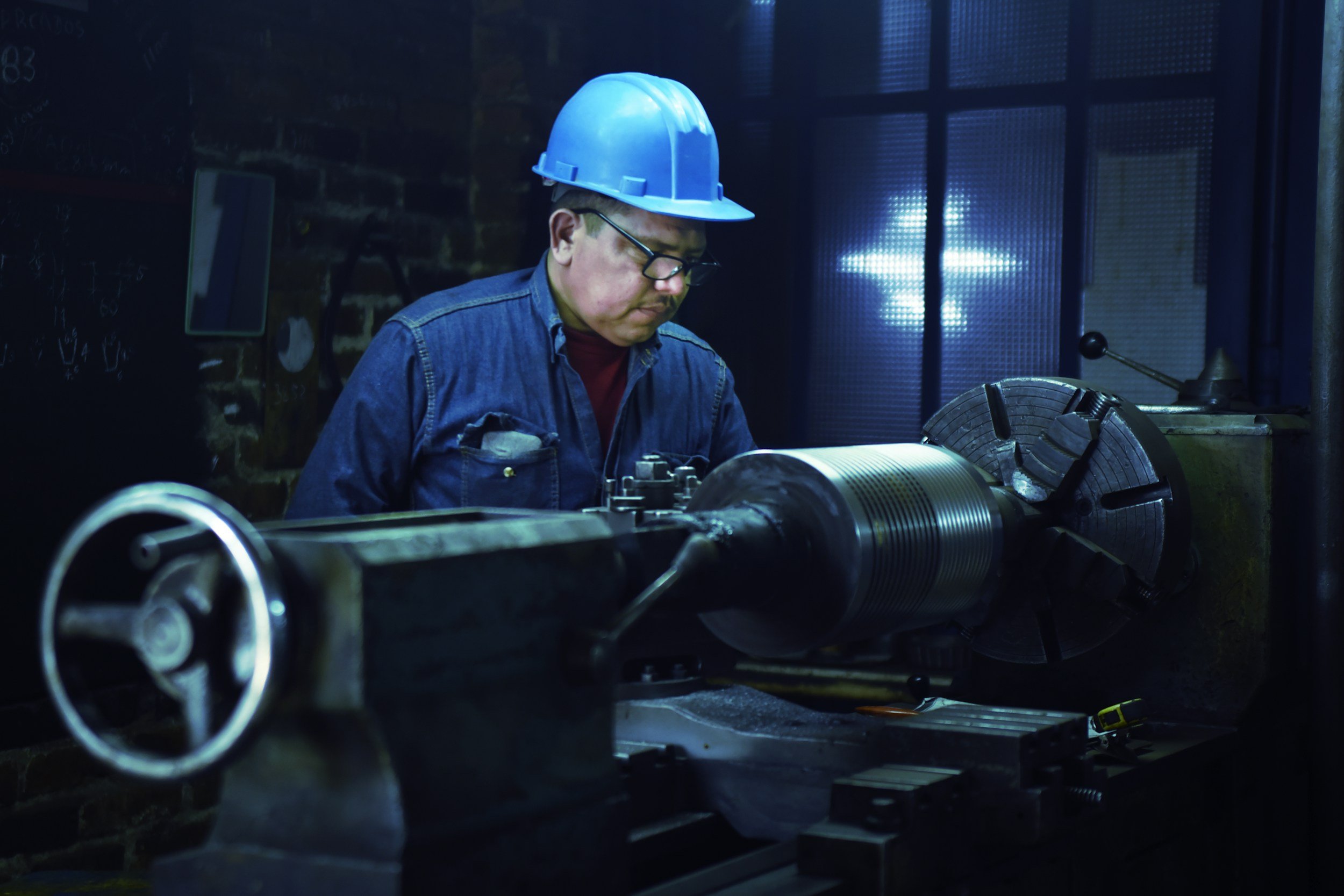 A man in a blue hard hat and glasses working on a piece of machinery in a dimly lit industrial setting.