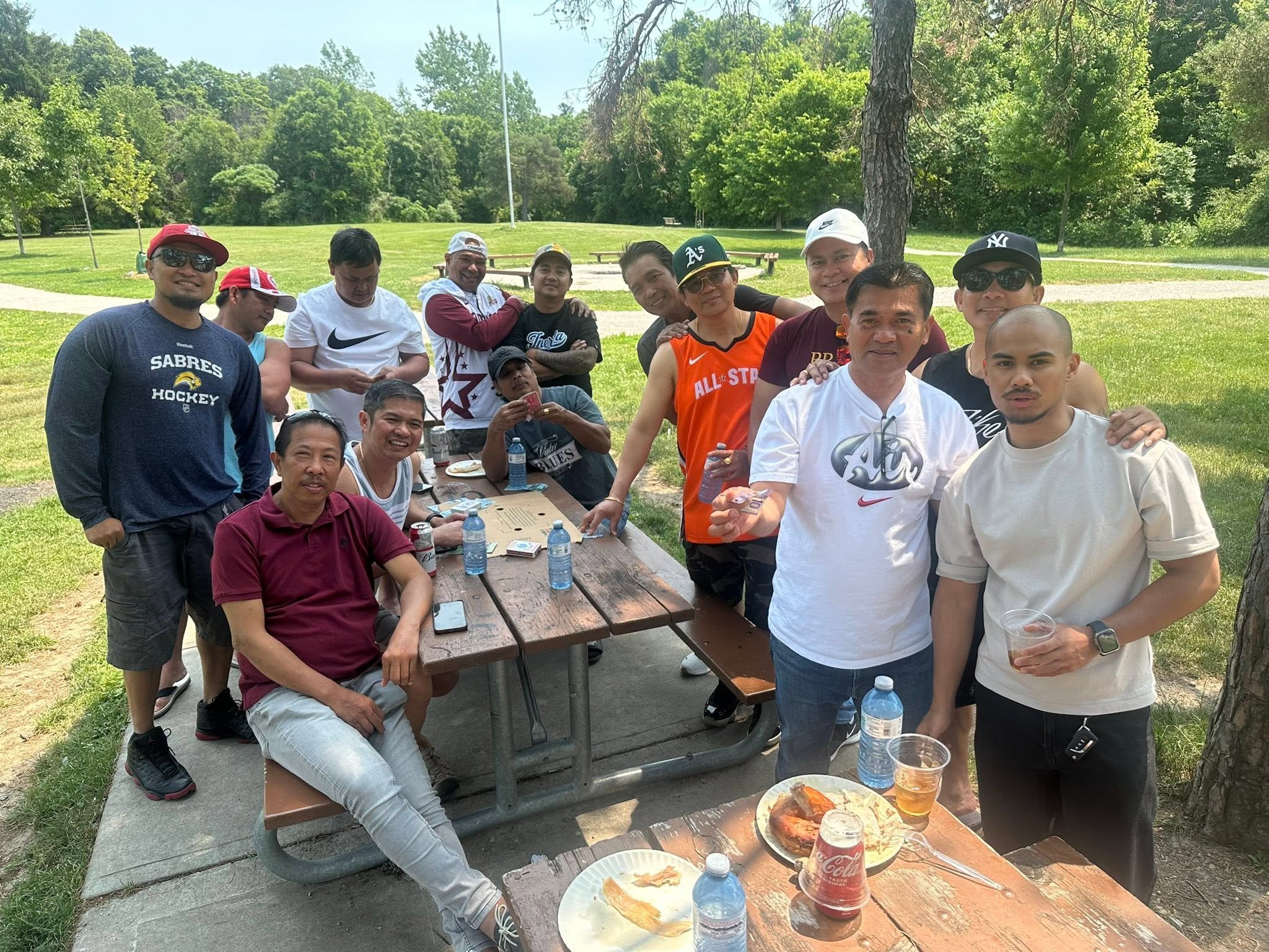 A group of people at a park during daytime, smiling and posing for a photo around a picnic table with food and drinks, with green trees and grass in the background.