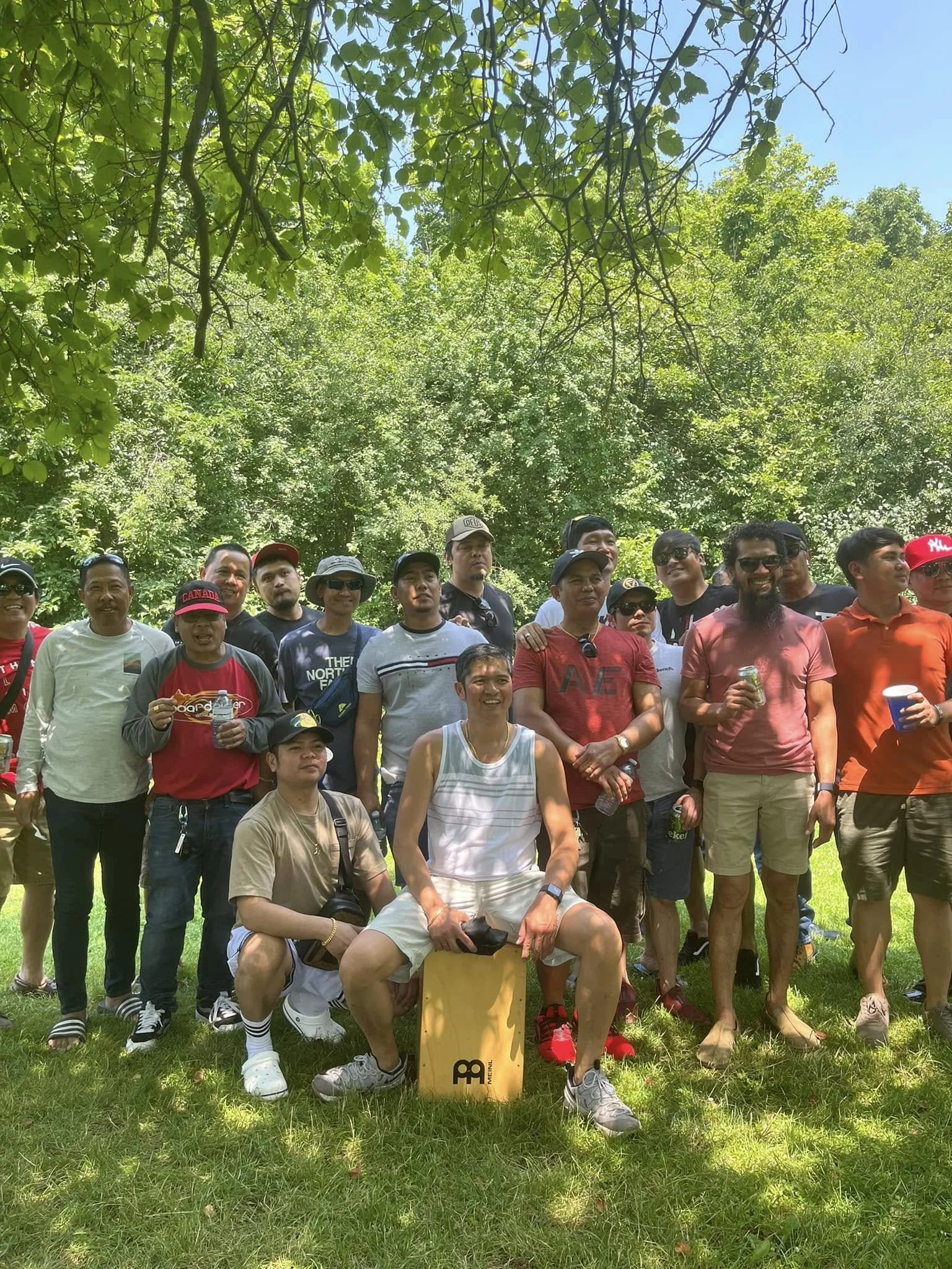 A group of people gathered outdoors in a park with green trees and blue sky overhead. They are smiling and some are holding drinks. In the front, a man is seated on a large, yellow block with the number 3 on it, wearing headphones around his neck, white socks with black stripes, and athletic shoes. The group appears to be enjoying a sunny day together.