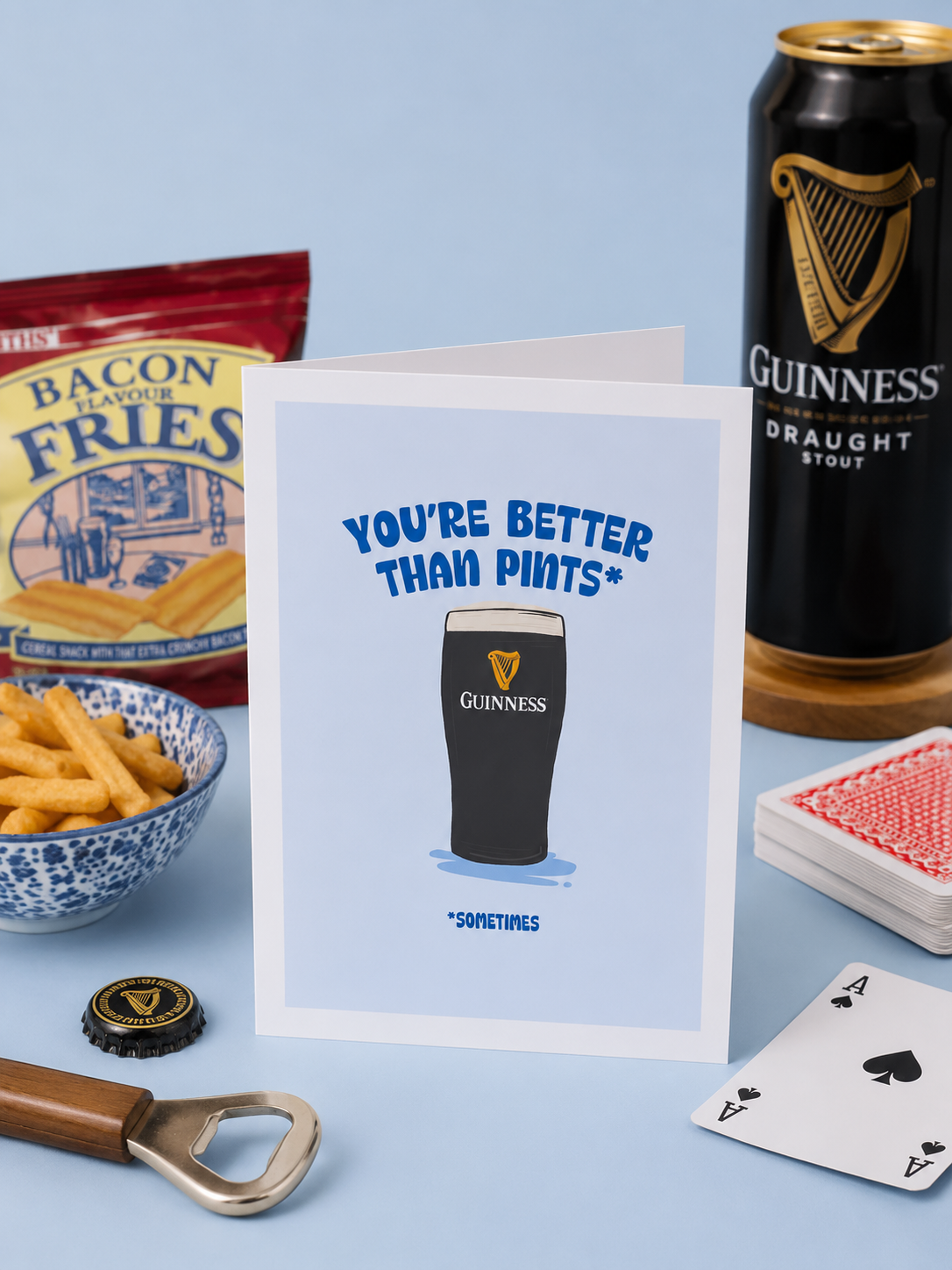 A greeting card with the message 'You're better than pints*' and a drawing of a Guinness pint glass. The card is surrounded by a bowl of fries, a bag of bacon-flavored fries, a can of Guinness Draught stout, a bottle opener, an ace of spades card, a black Guinness bottle cap, and a deck of cards on a light blue surface.