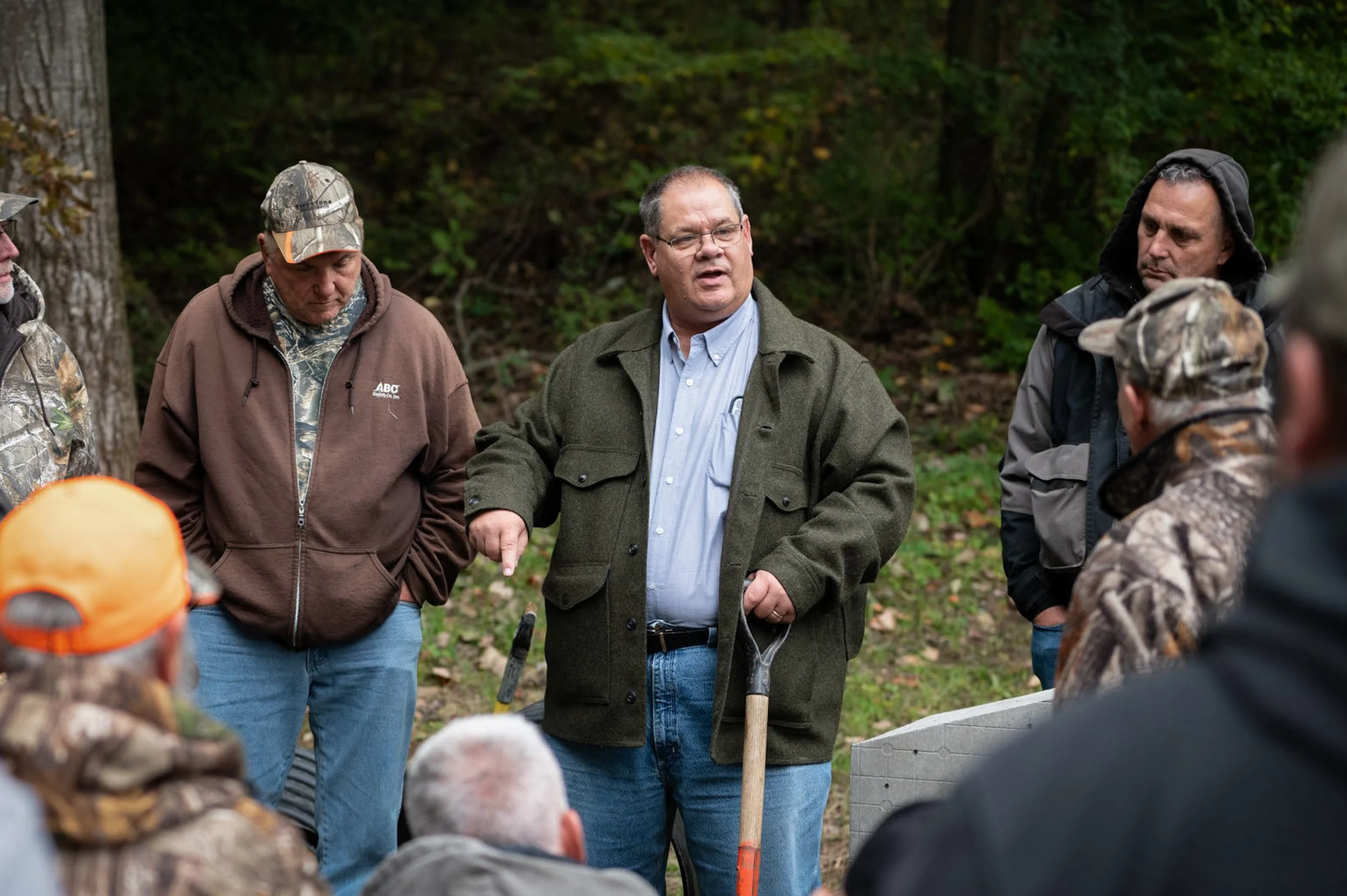 A group of people gathered outdoors in a wooded area, with a man in glasses and a green jacket speaking to the group, while others listen.
