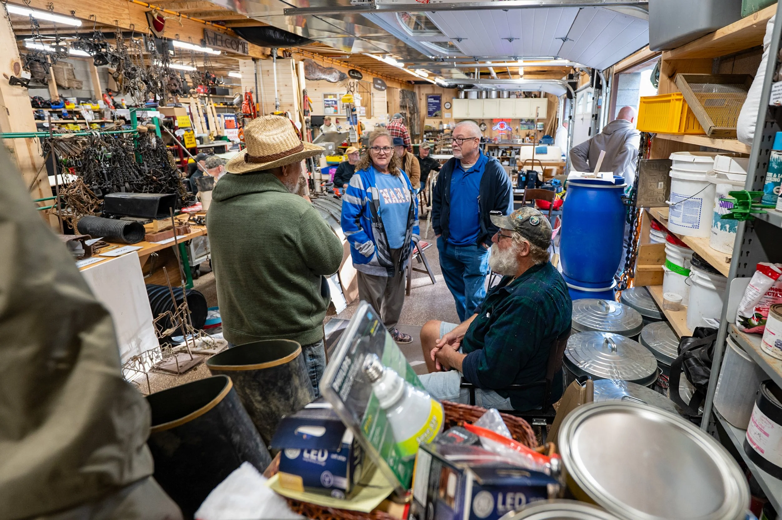 People inside a cluttered hardware or antique store with shelves filled with various tools, containers, and miscellaneous items. Several individuals are engaged in conversation, some standing and one sitting on a stool, surrounded by vintage and hardware supplies.
