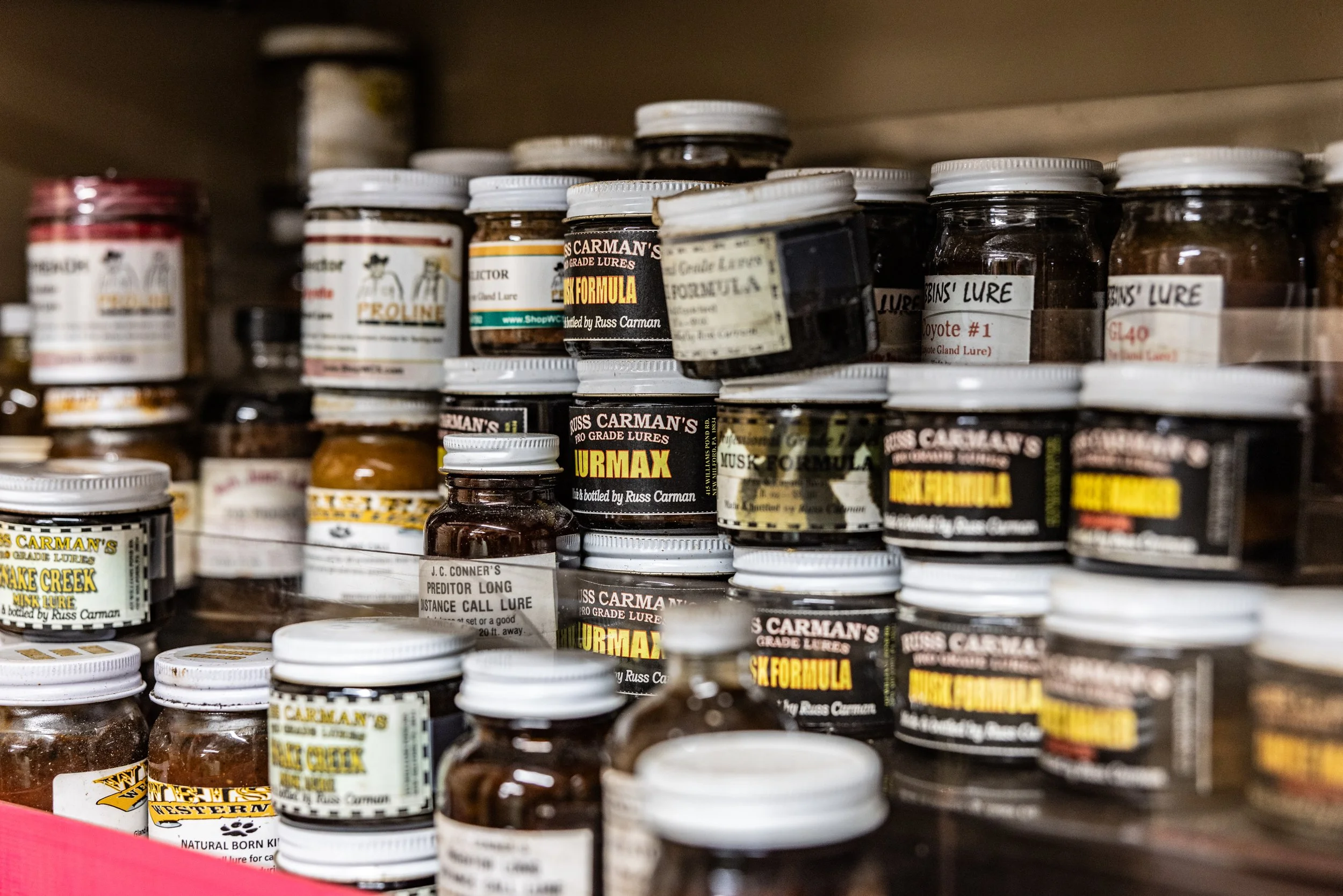 A collection of small jars of fishing bait or lures on shelves in a store. The labels include brand names and lure descriptions, some with handwritten notes.