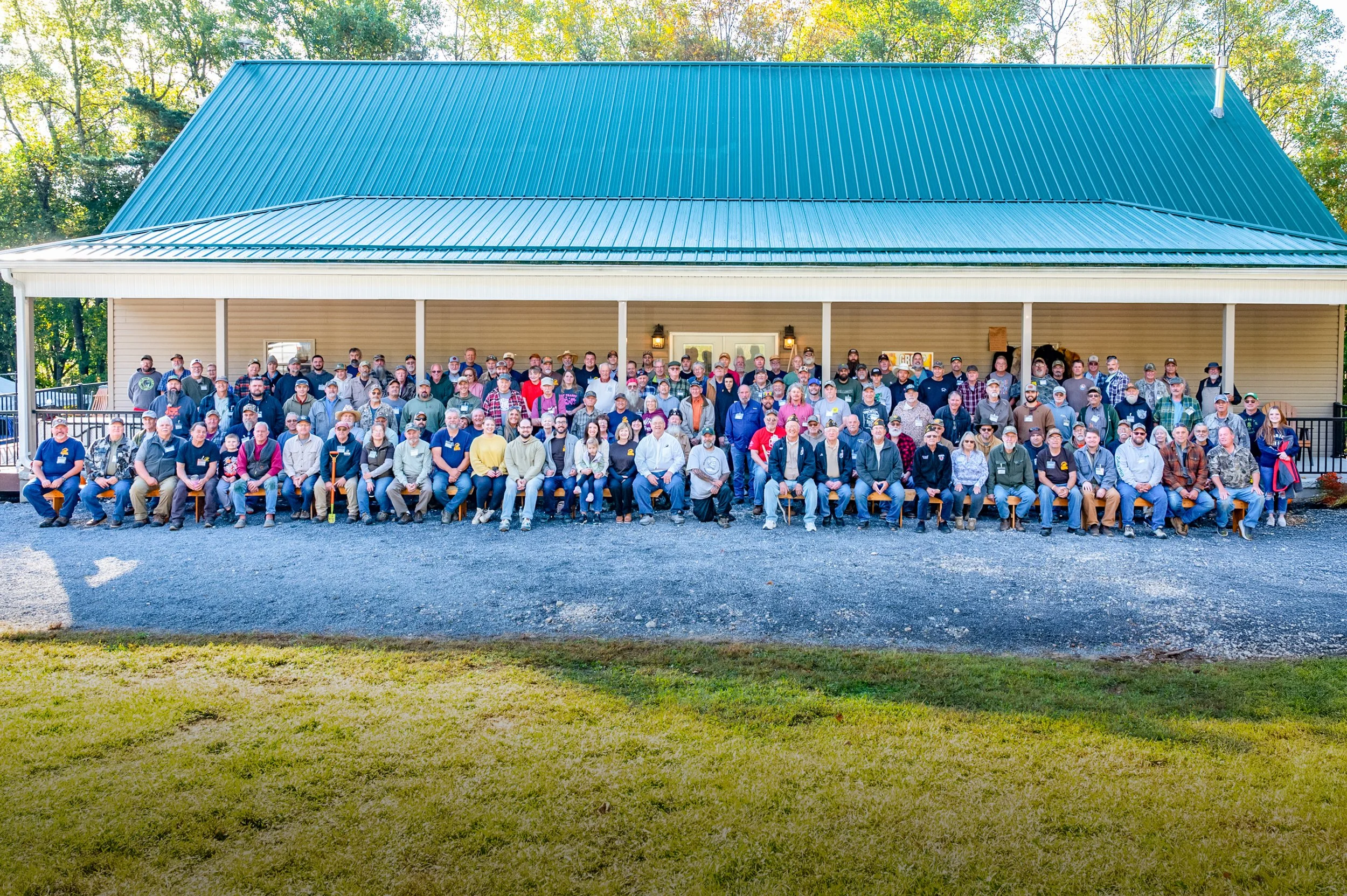 A large group of people gathered outside in front of a house with a teal metal roof and beige siding, during daylight.