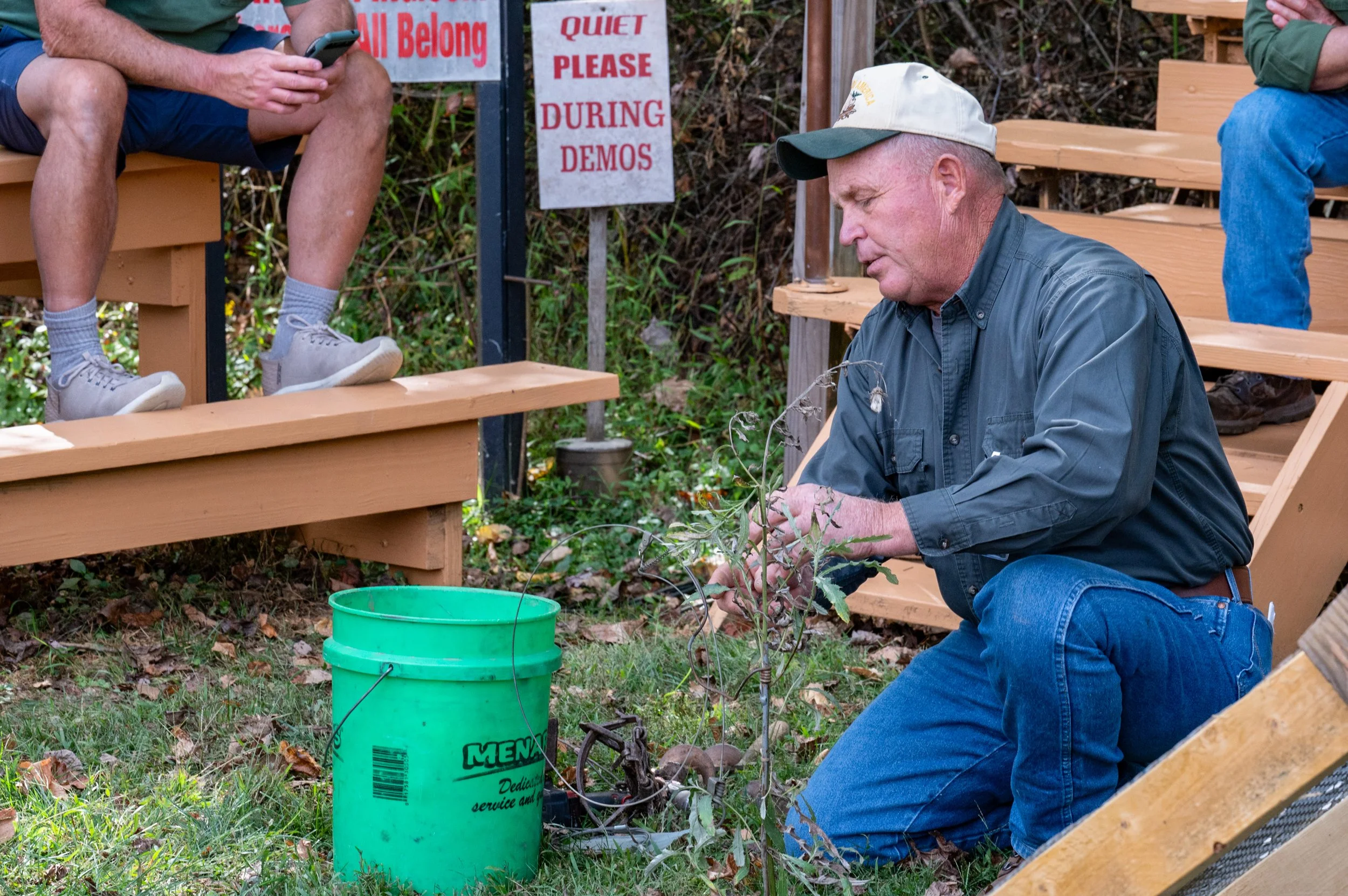 A man kneeling outdoors planting a small tree or shrub, with two other people seated on benches nearby, possibly during a community event or demonstration. There are signs in the background that read 'Quiet Please During Demos' and 'All Belong'.
