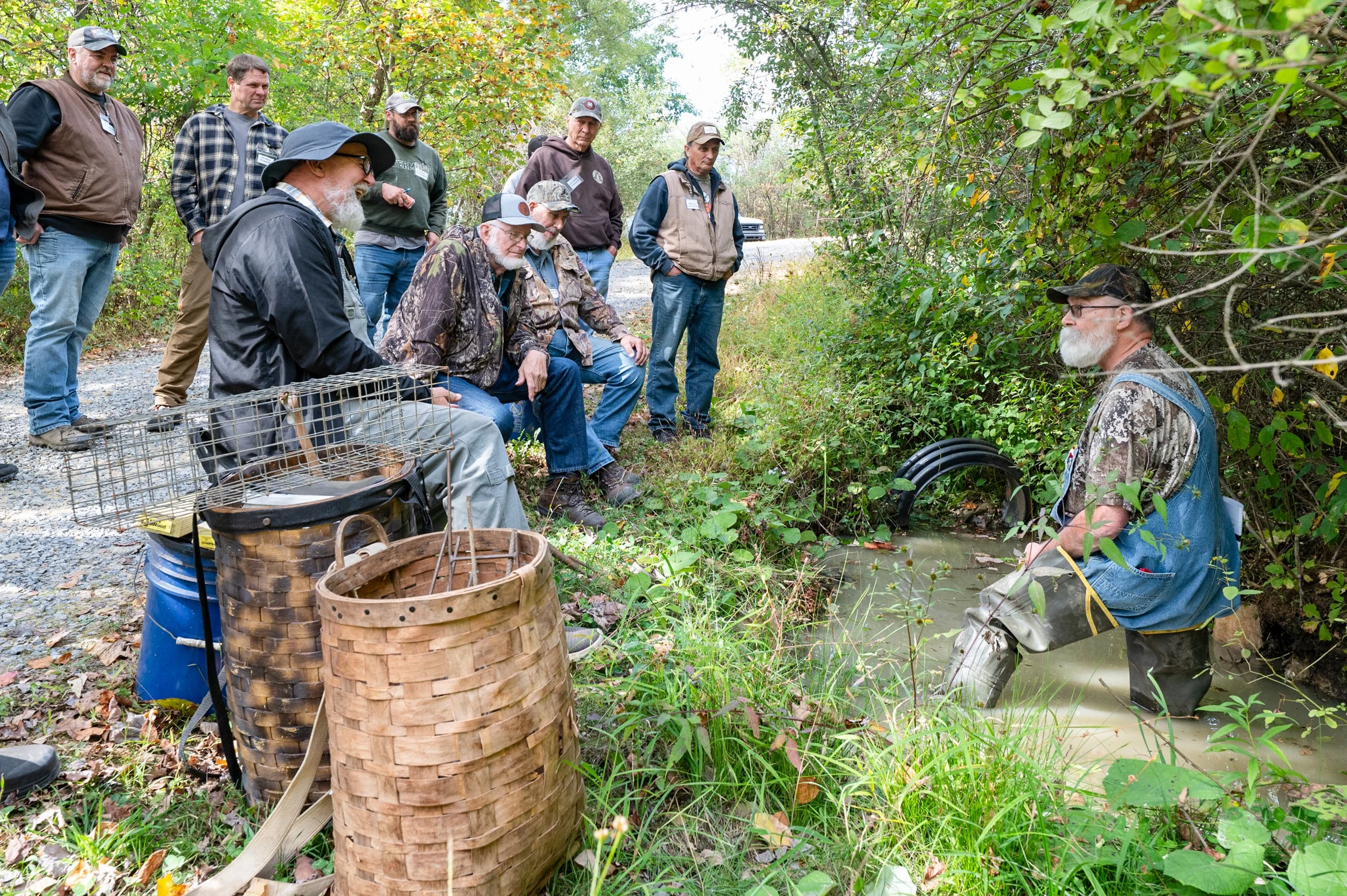 Group of people gathered outdoors near a small stream, with one person kneeling in the water, wearing camouflage waders, while others stand and sit nearby, surrounded by trees and foliage.