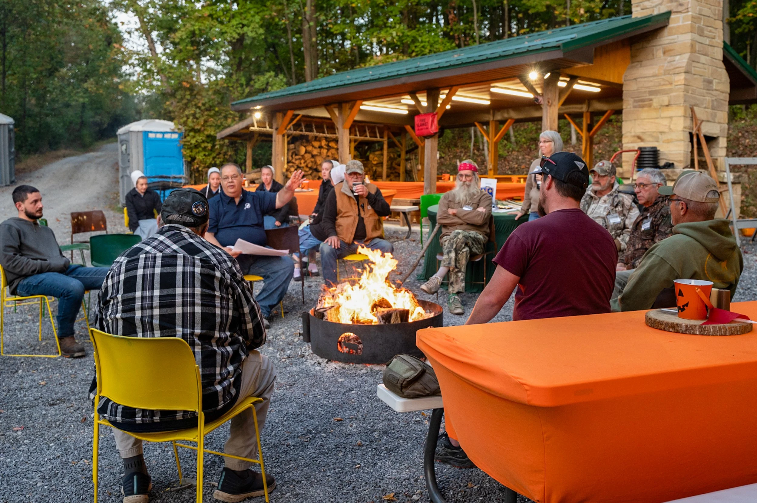 A group of people gathered outdoors around a campfire during evening, some sitting on colorful chairs, with a covered shelter, trees, and a gravel path in the background.