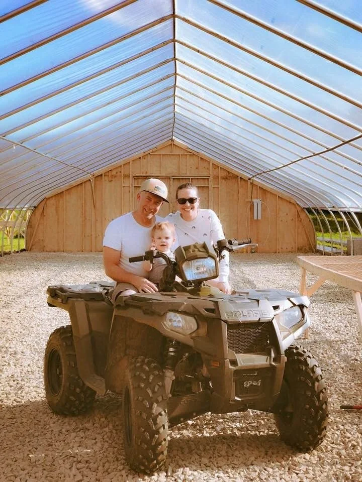 Family with a young girl sitting on a toy all-terrain vehicle inside a large greenhouse with wooden walls and a transparent arched roof.
