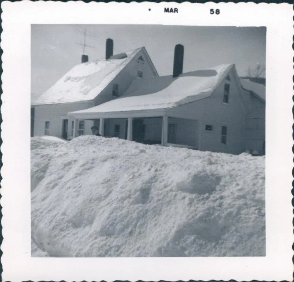 A black-and-white photo of a house covered in snow, with large snowbanks in the foreground and snow on the roof, taken in March 1958.
