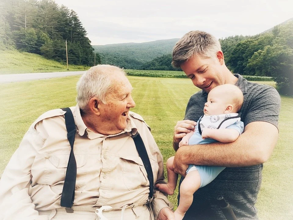 A young man holding a baby in a field, smiling at an elderly man, in a green outdoor landscape.