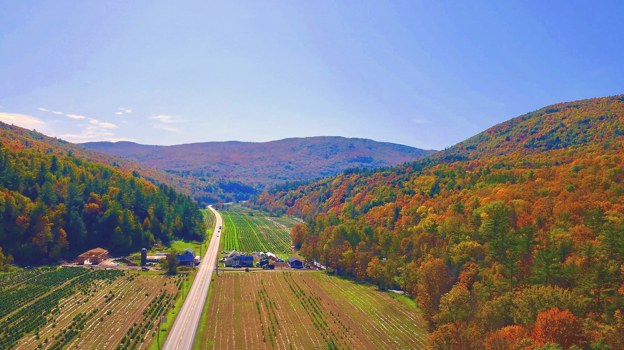 A scenic view of a valley with farmland and a small road, surrounded by colorful autumn trees on rolling hills and mountains under a clear blue sky.