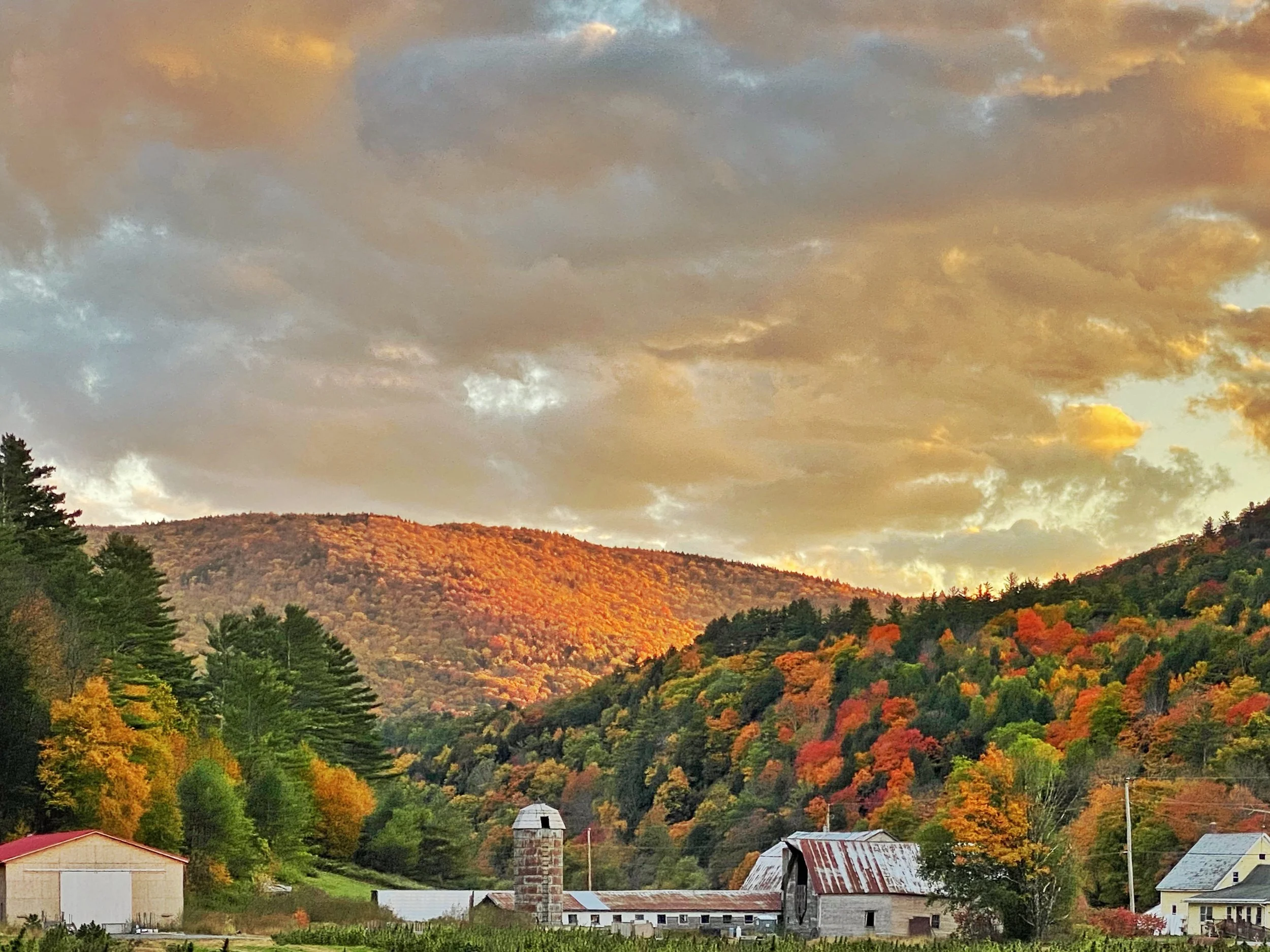 A scenic view of a rural area with farm buildings, trees, and rolling hills covered in fall foliage during sunset, with a cloudy sky overhead.