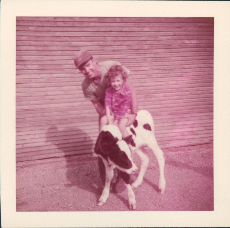 A man and a young girl riding a calf in front of a wooden building.