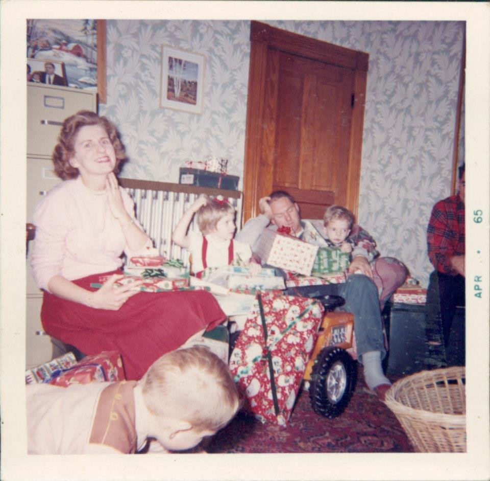 Family gathered around during Christmas, children opening presents, adults sitting on a couch with wrapped gifts, decorated for the holiday season.