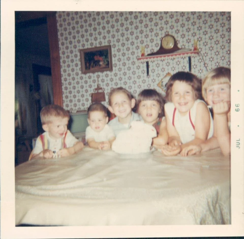 Six children sitting around a table, celebrating a birthday with a cake, in a vintage kitchen with patterned wallpaper, a clock, and a shelf with decorations in the background.