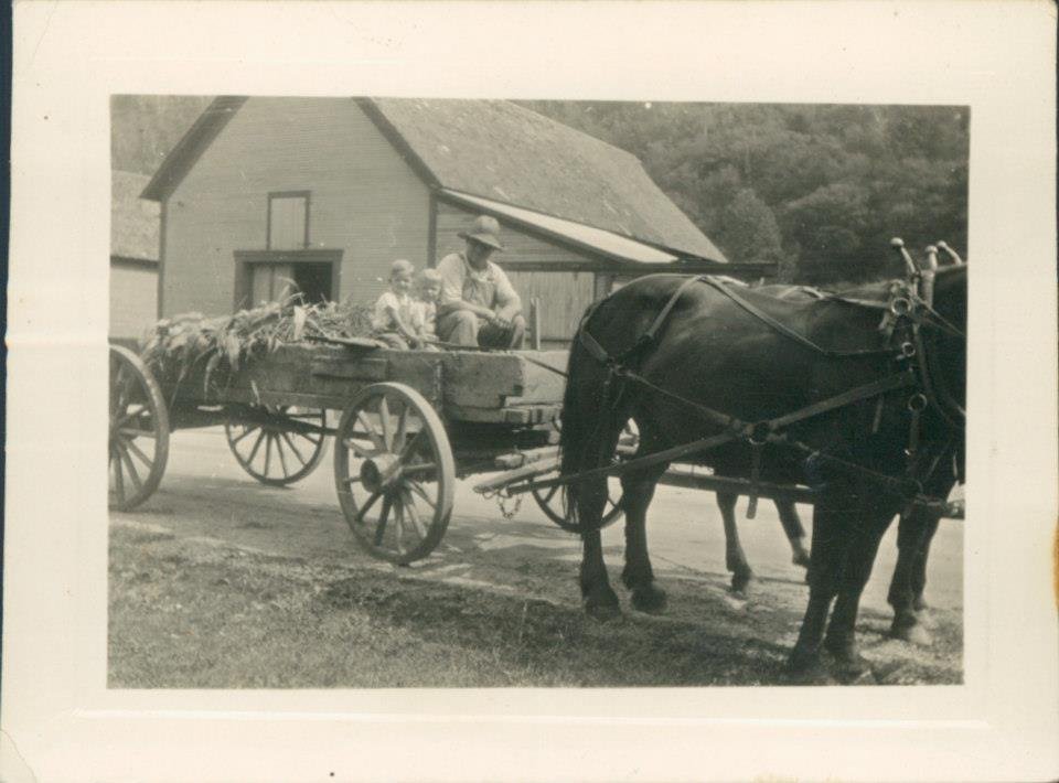 Black and white photo of two people sitting in a horse-drawn wagon filled with hay, pulled by a horse, in front of a barn or farmhouse.