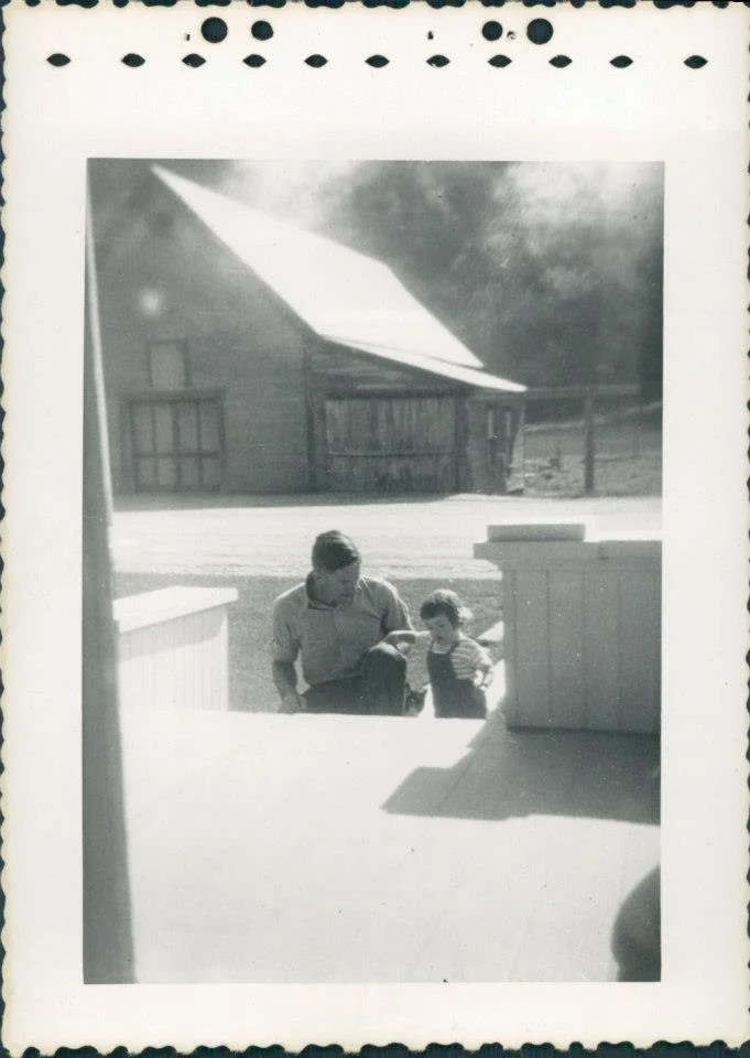 A black and white photo of a man and a young girl standing outside on a porch or deck area, with a building and a shed in the background. The man is bending down and looking at the girl.