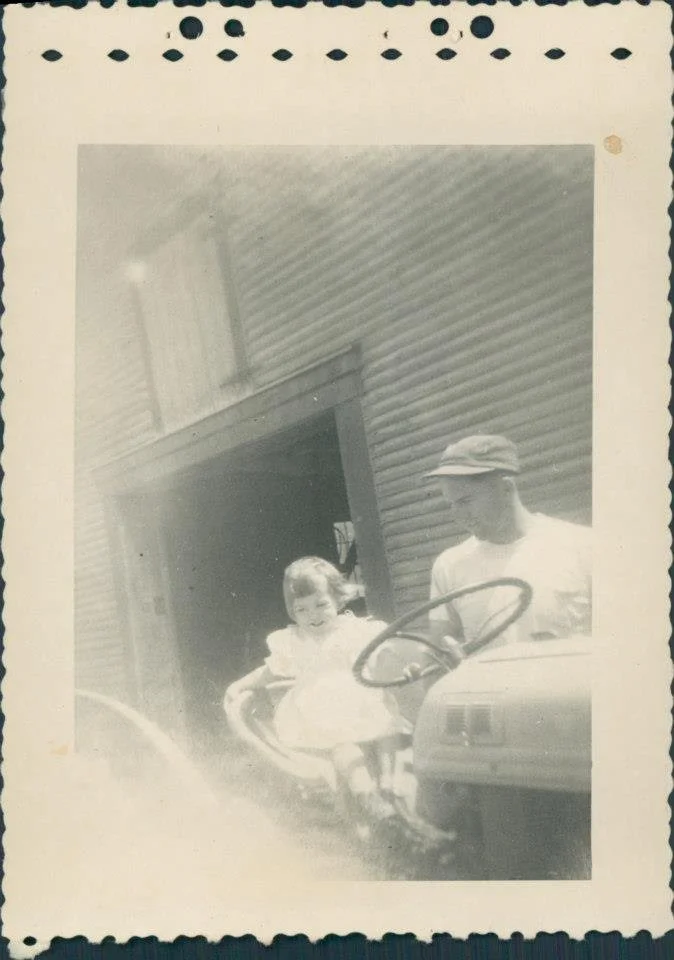 A black-and-white photograph of a young girl sitting on a tractor, smiling at the camera, with a man, possibly her father, standing beside her. They are outside a house with a wooden exterior.