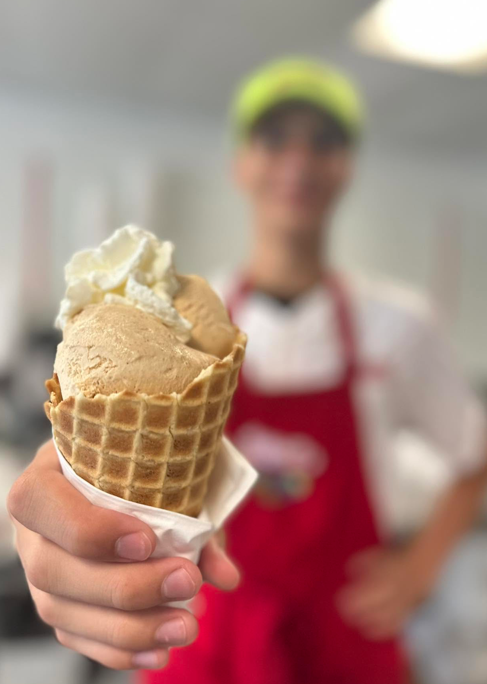 Person holding a waffle cone with two scoops of vanilla ice cream, with a blurred person in a red apron and yellow hat in the background.