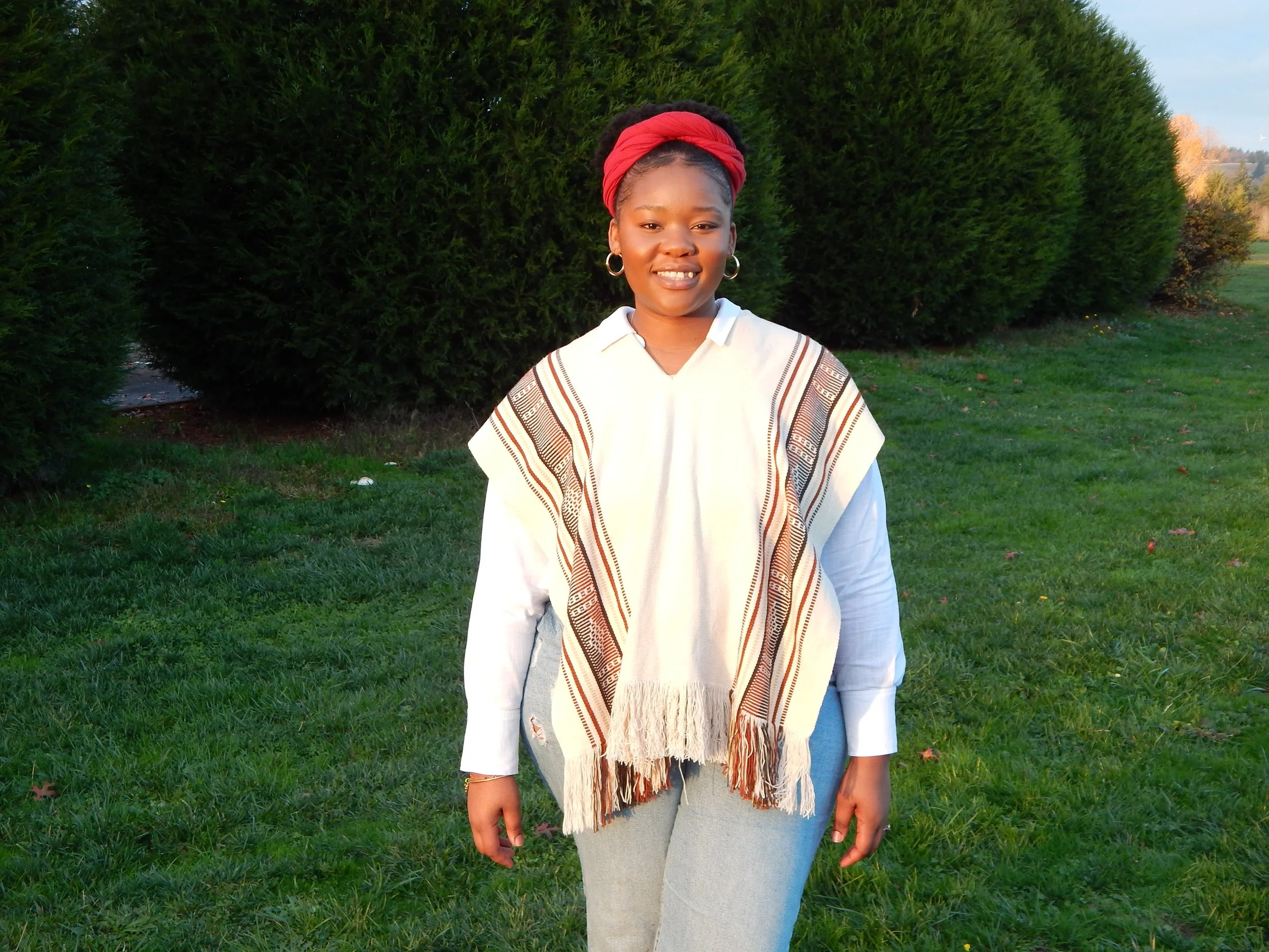 A smiling woman standing on grass with a large green hedge in the background, dressed in casual clothing, wearing hoop earrings, and a red headscarf.