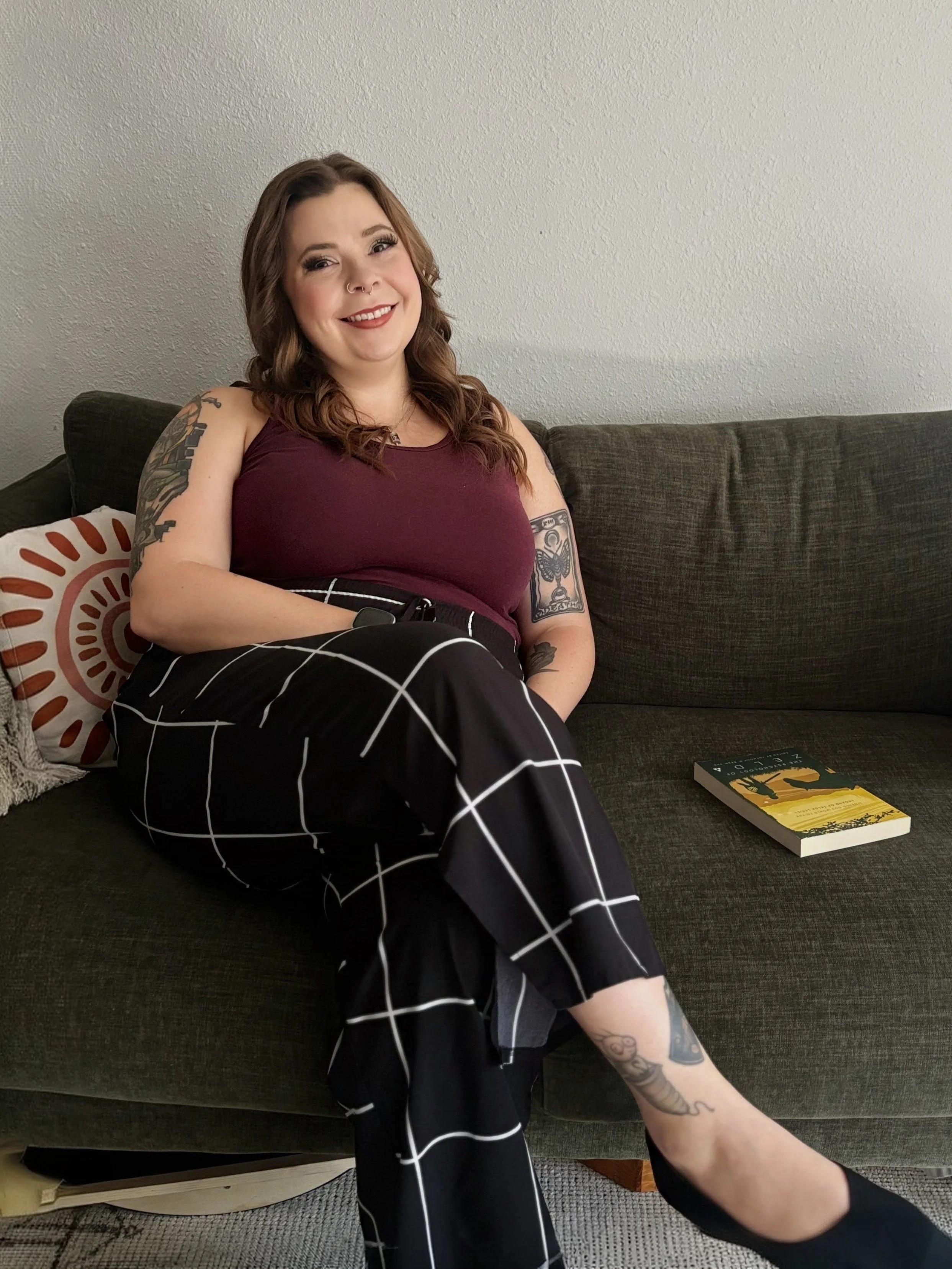 A woman with brown hair, tattoos, and a septum piercing sitting on a dark sofa, smiling, wearing a burgundy top and black and white checkered pants, with a book on the sofa nearby.