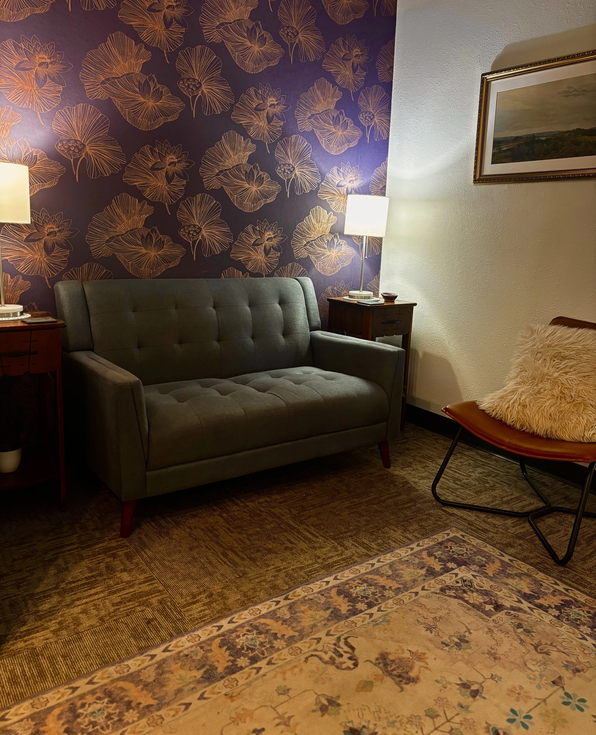 A cozy living room corner with a green tufted sofa, a wooden side table, a white lampshade lamp, a framed landscape painting on the white textured wall, and a patterned rug covering the floor. The background features a dark purple wallpaper with outlined floral patterns.
