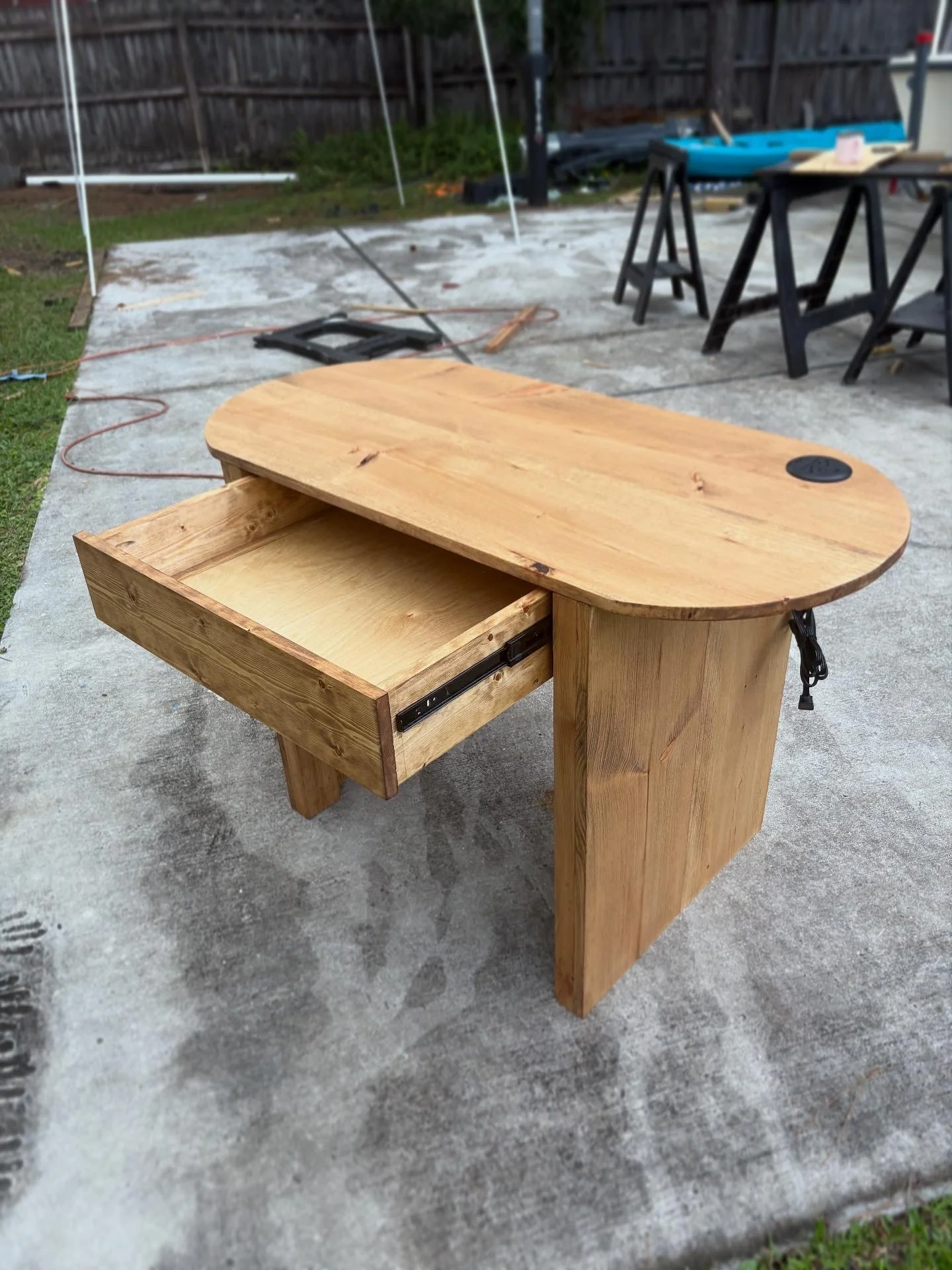 Unfinished wooden desk with a single drawer and black metal slides, set outdoors on a concrete surface.
