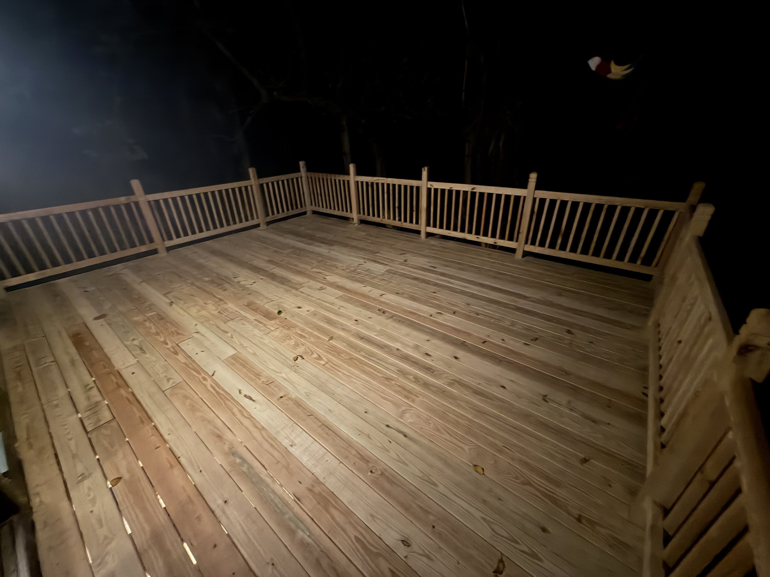 Empty wooden deck with railing at night, surrounded by dark trees, and a small bird flying in the dark sky.