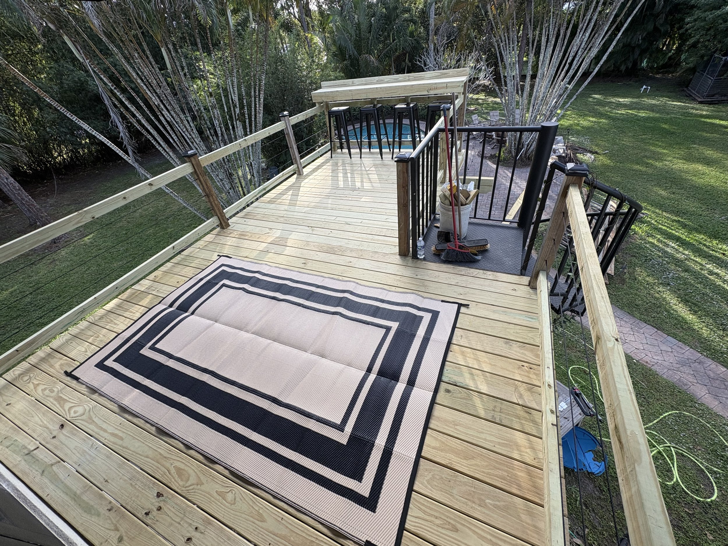 Outdoor wooden deck with black metal railings, a black and white striped rug, cleaning supplies, and a small blue pool in a backyard with green grass and trees.