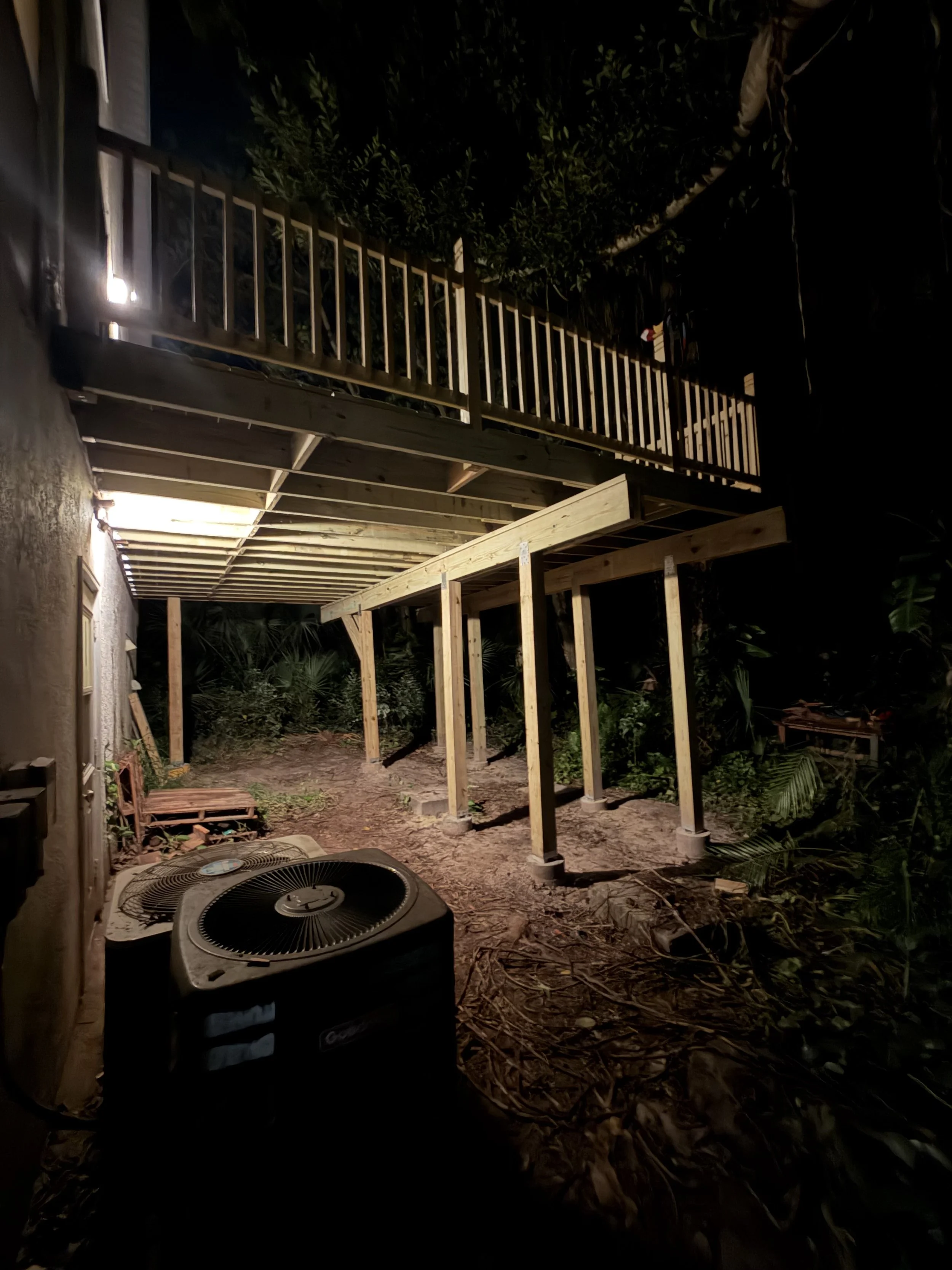 Nighttime backyard scene with an incomplete wooden deck; the frame structure and raised platform are under construction, surrounded by plants and leaves, with an air conditioning unit in the foreground and a staircase railing visible.