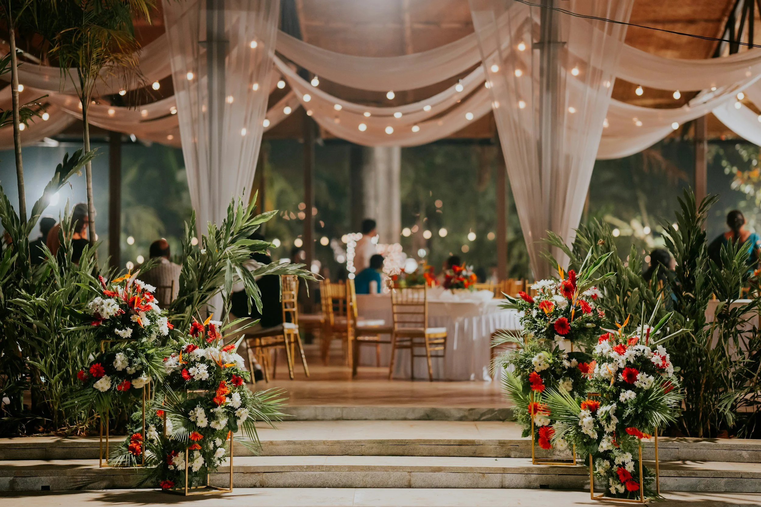 An indoor wedding venue decorated with white drapes and string lights, with tables, chairs, and floral arrangements featuring red, white, and orange flowers, and some guests seated and standing in the background.