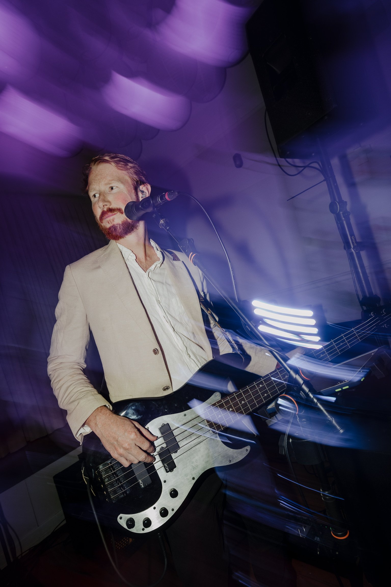 A man with a beard wearing a beige blazer playing an electric bass guitar while singing into a microphone in a dimly lit room with purple lighting.