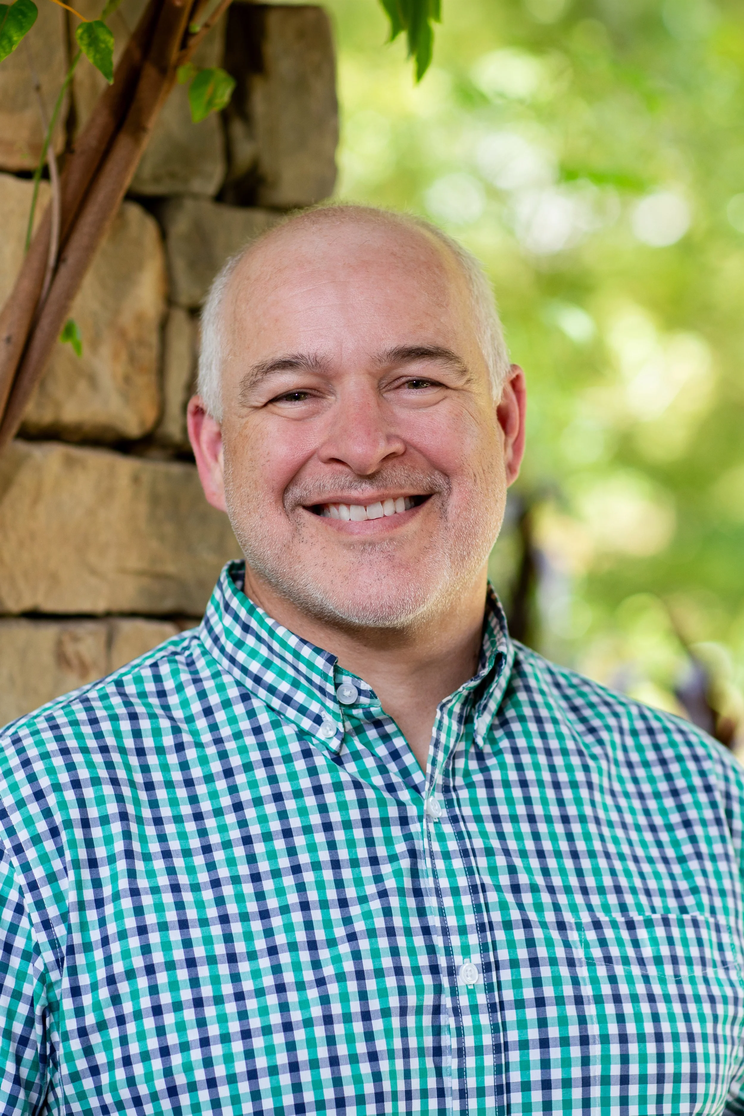 A smiling middle-aged man with short, light gray hair wearing a green and blue checkered shirt. Background is blurred with green foliage and a stone wall.