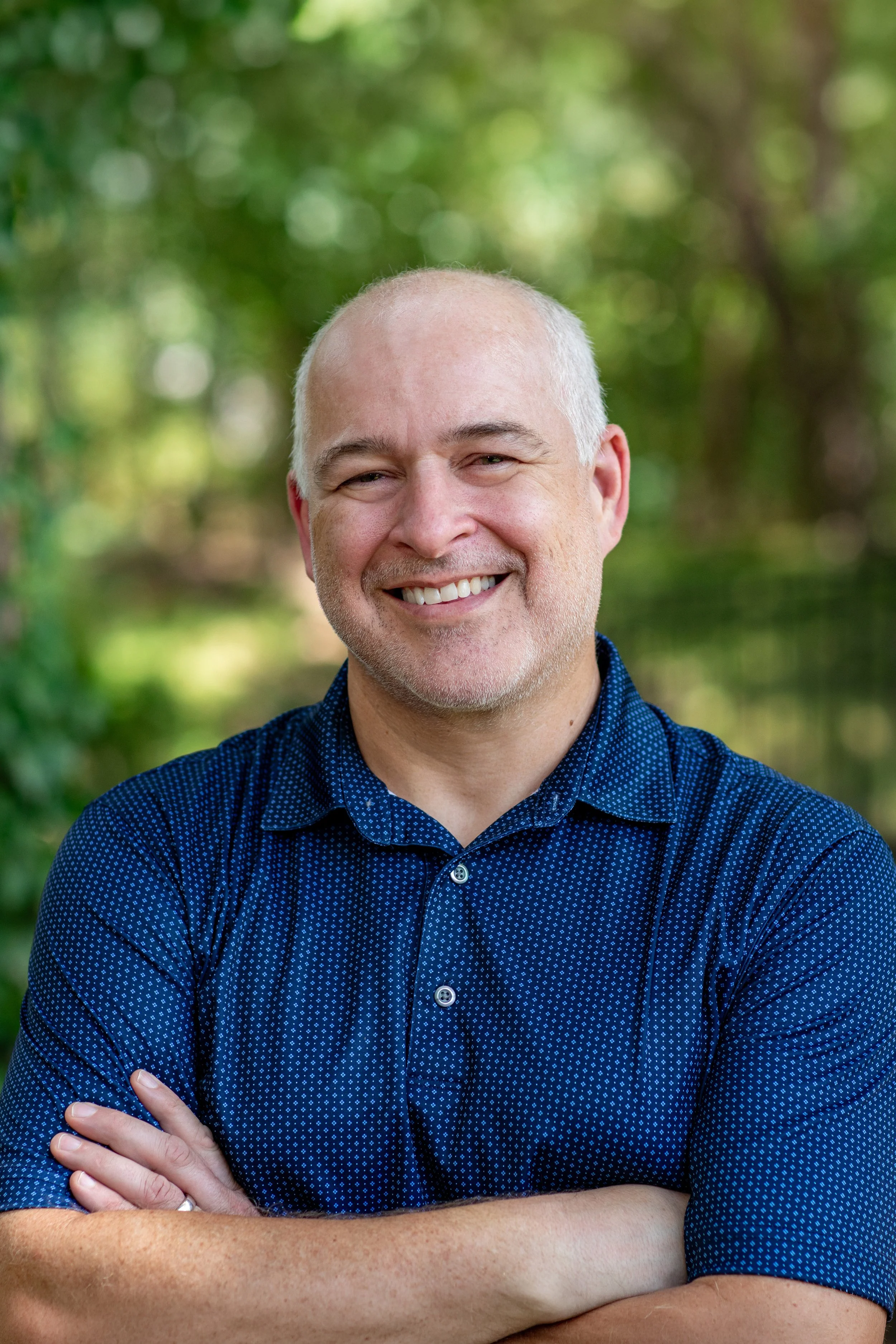 A smiling middle-aged man with a bald head, wearing a blue patterned button-up shirt, standing outdoors with greenery in the background.