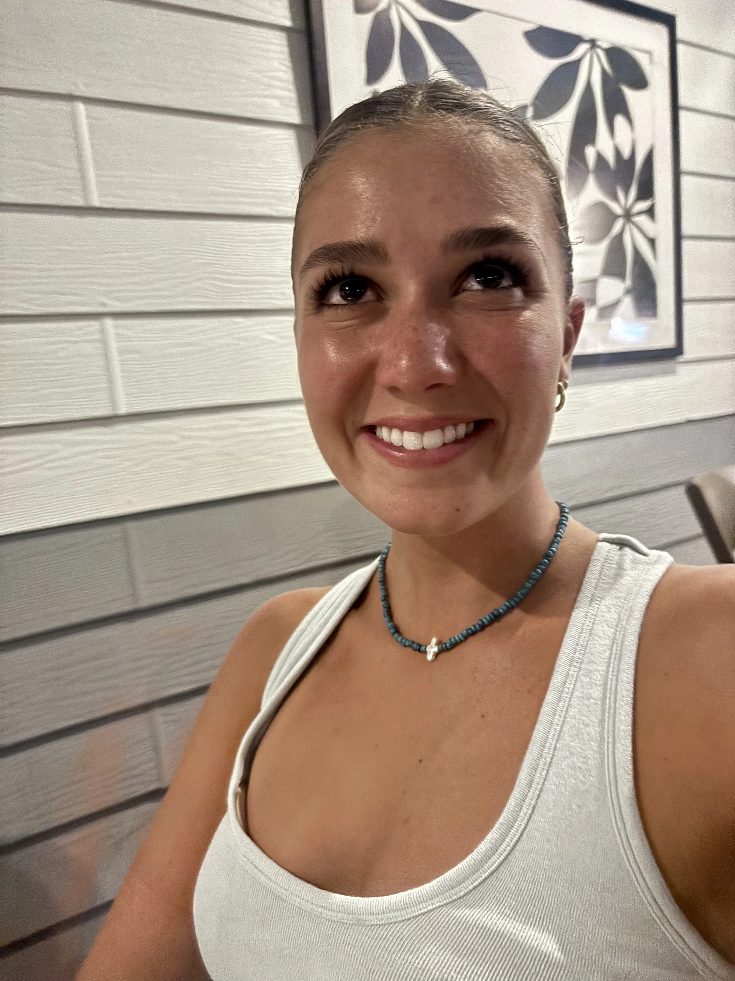 A young woman wearing a white tank top, blue beaded necklace, and earrings, smiling in front of a white and gray wall with a framed black-and-white floral art piece.