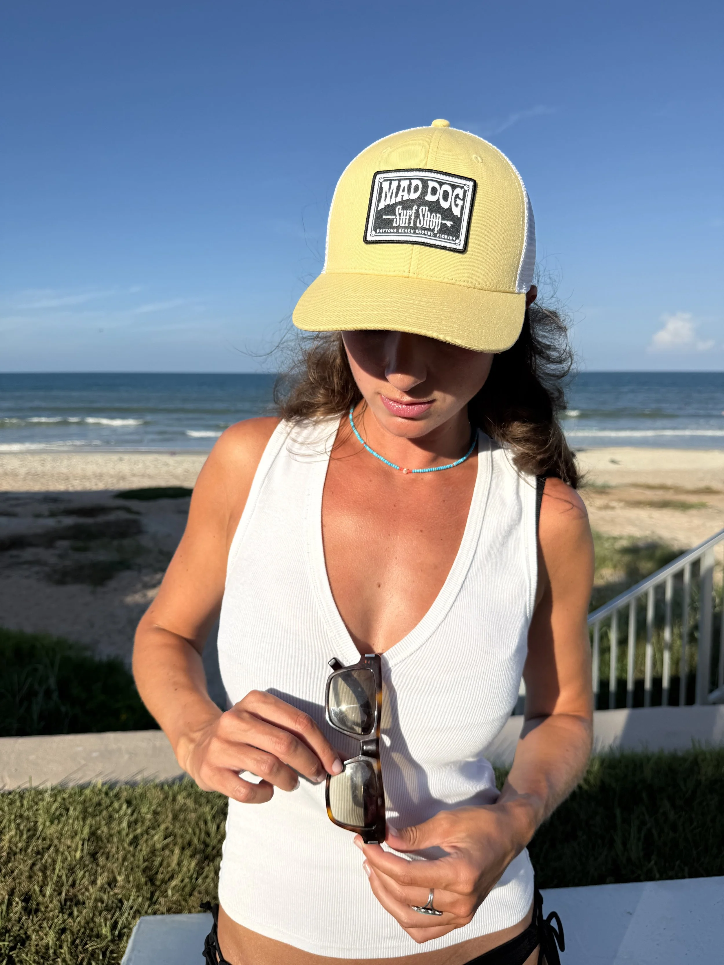 A woman wearing a yellow and white baseball cap with a "Mad Dog Surf Shop" patch logo, standing at the beach with the ocean and clear blue sky in the background, holding sunglasses and wearing a white tank top.