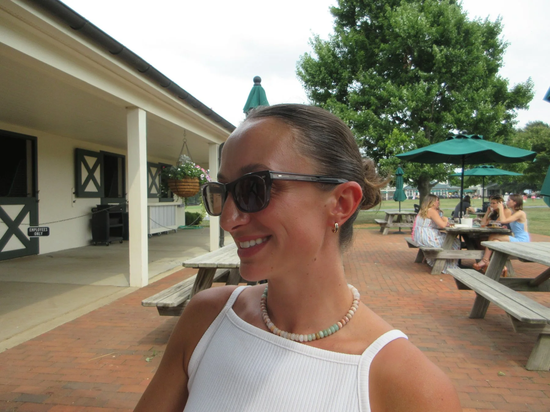 A woman wearing sunglasses, a white sleeveless top, and a pearl necklace smiling outdoors at a park or outdoor dining area.