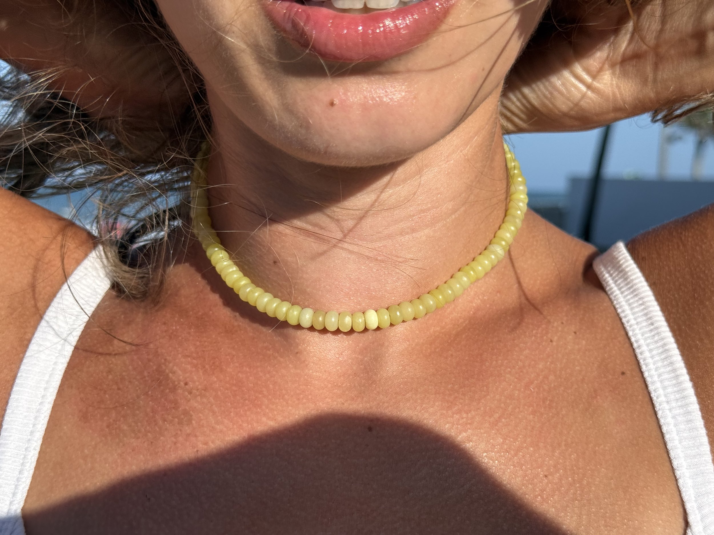 Close-up of a woman’s lips, chin, and neck wearing a yellow gemstone-beaded necklace and a white sleeveless top, with sunlight and a beach background.