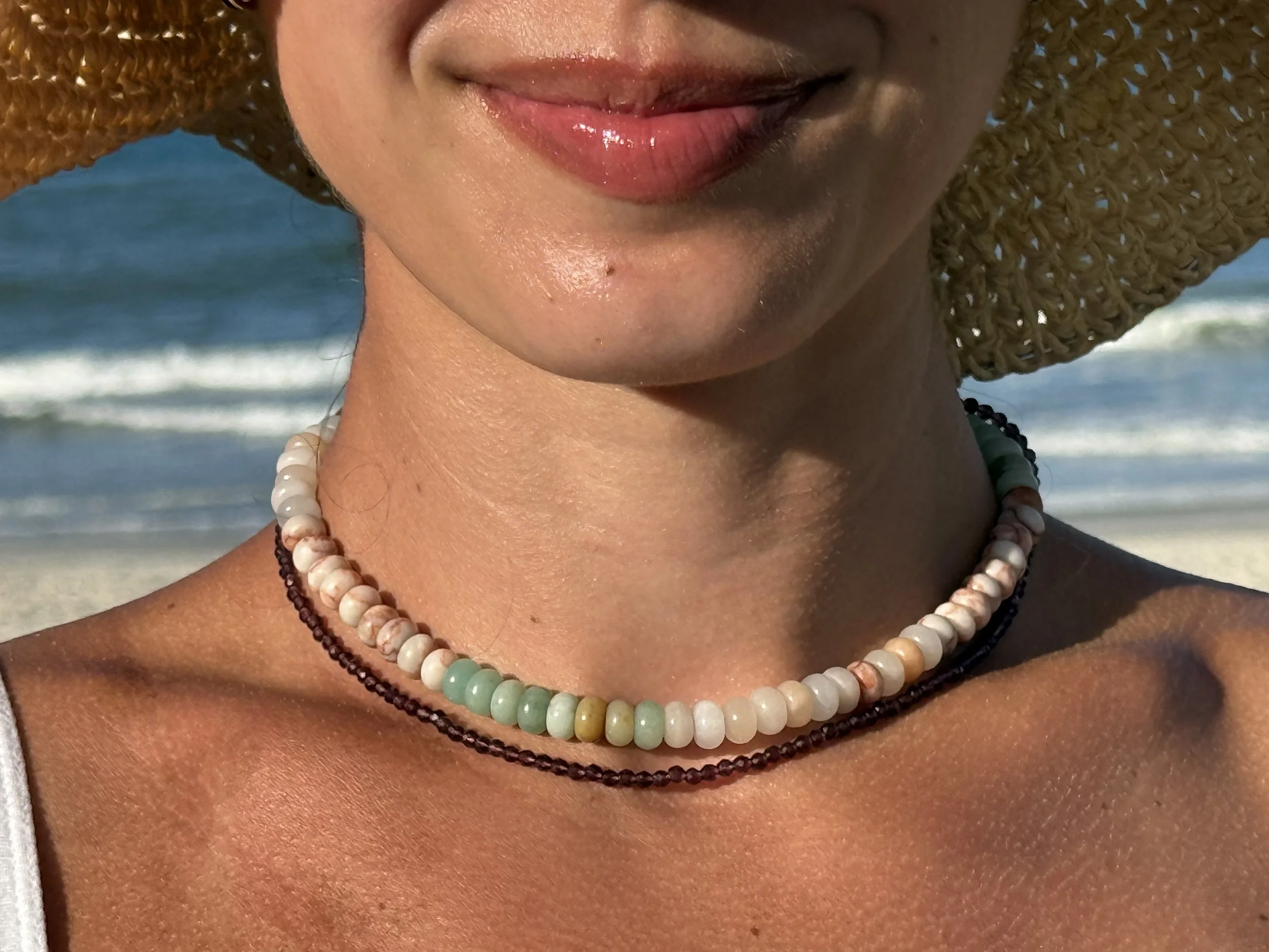 Close-up of a woman smiling with lipstick, wearing a wide-brimmed straw hat, layered beaded necklaces, and sitting near the ocean with waves in the background.