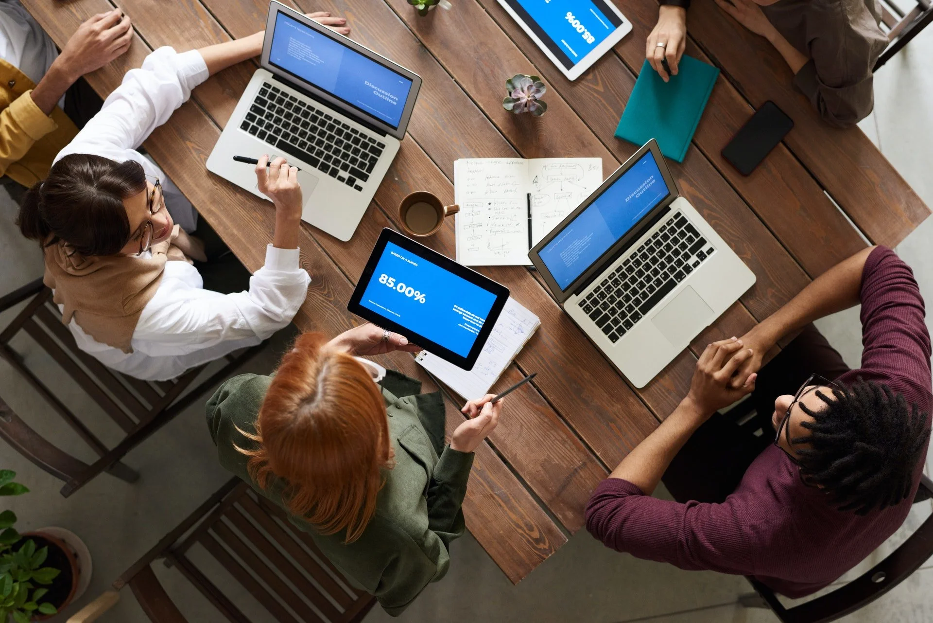Top-down view of a diverse group of people collaborating at a wooden table, with laptops, tablets, notebooks, a coffee mug, and a small potted plant.