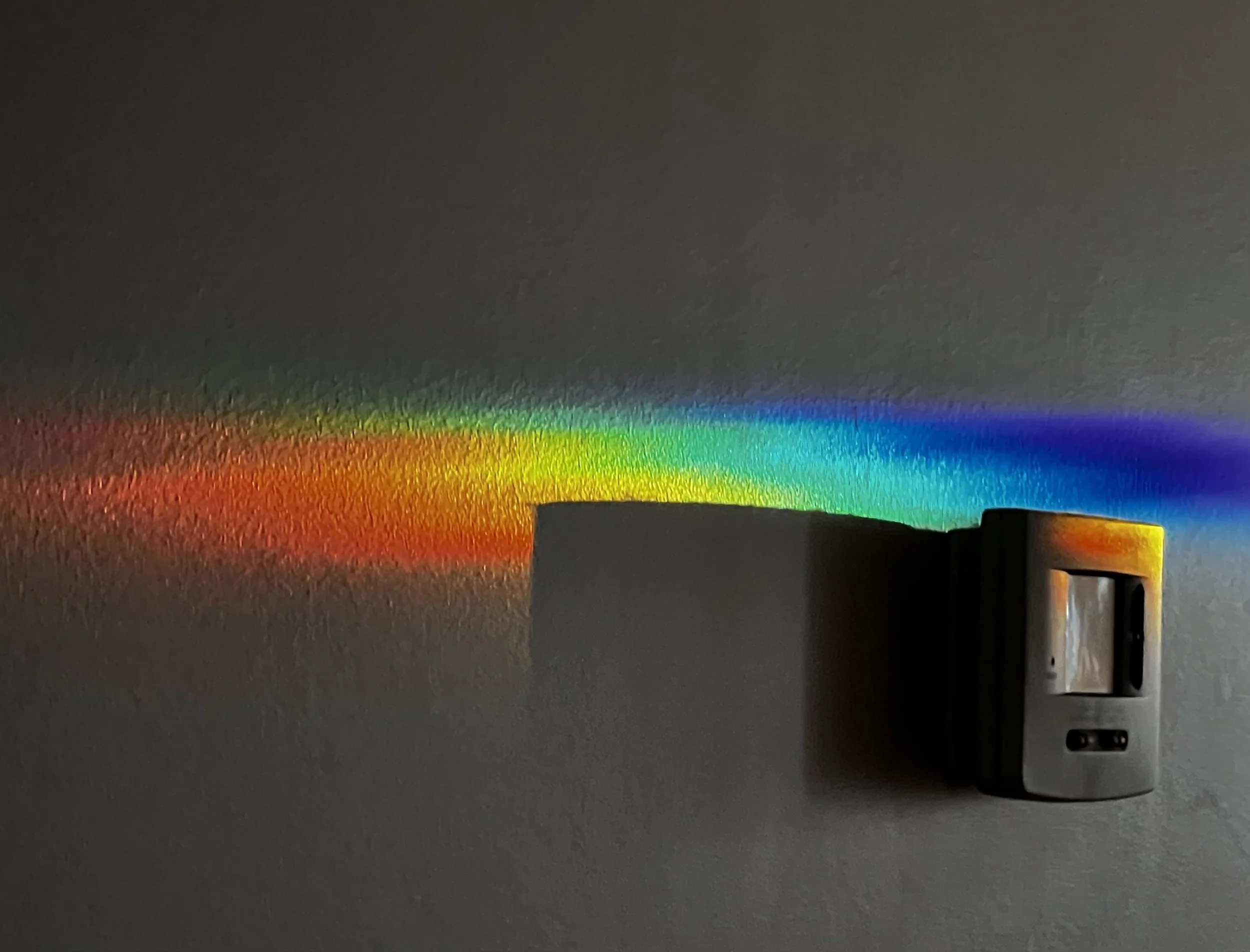 A rainbow spectrum visible on a dark gray wall, with the light source directed at a metallic object on the right side of the image.